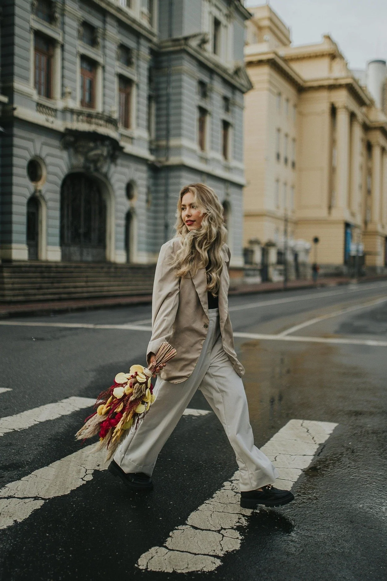 A woman walking across a city street crosswalk holding a bouquet of flowers, with historical buildings in the background.