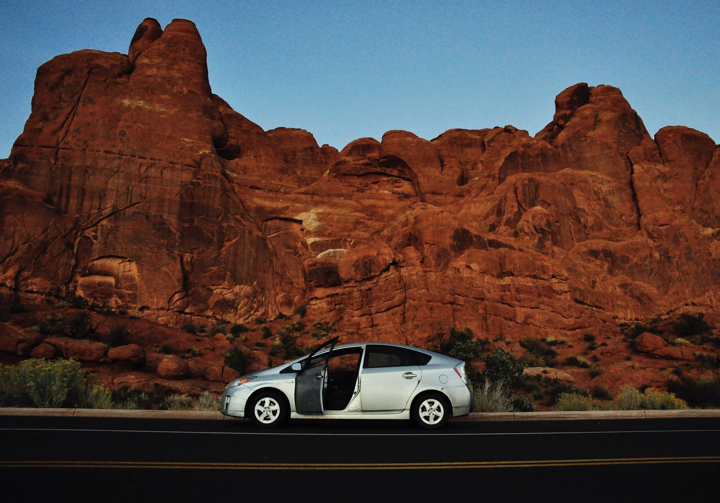 Photographer Kristin Godfrey's silver Prius with open driver's door parked on an empty road with large red rock formations in the background under a clear blue sky from roadtrip.