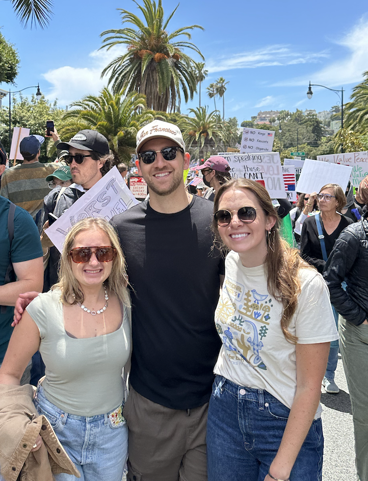 Solstice Studio's photographer Kristin Godfrey posing at a protest or rally, with signs and a crowd of protesters in the background, palm trees, and a clear blue sky.