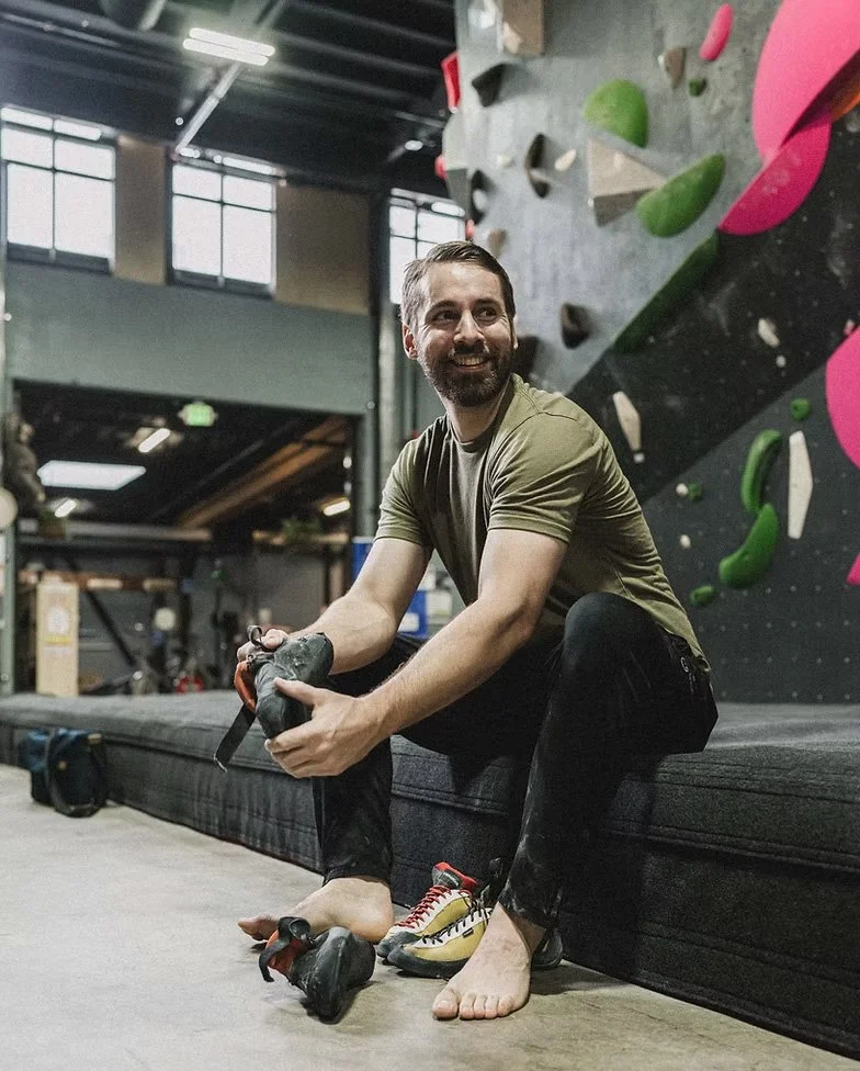 Man with a beard sitting on a rock climbing gym floor, smiling and putting on climbing shoes, with a climbing wall in the background.