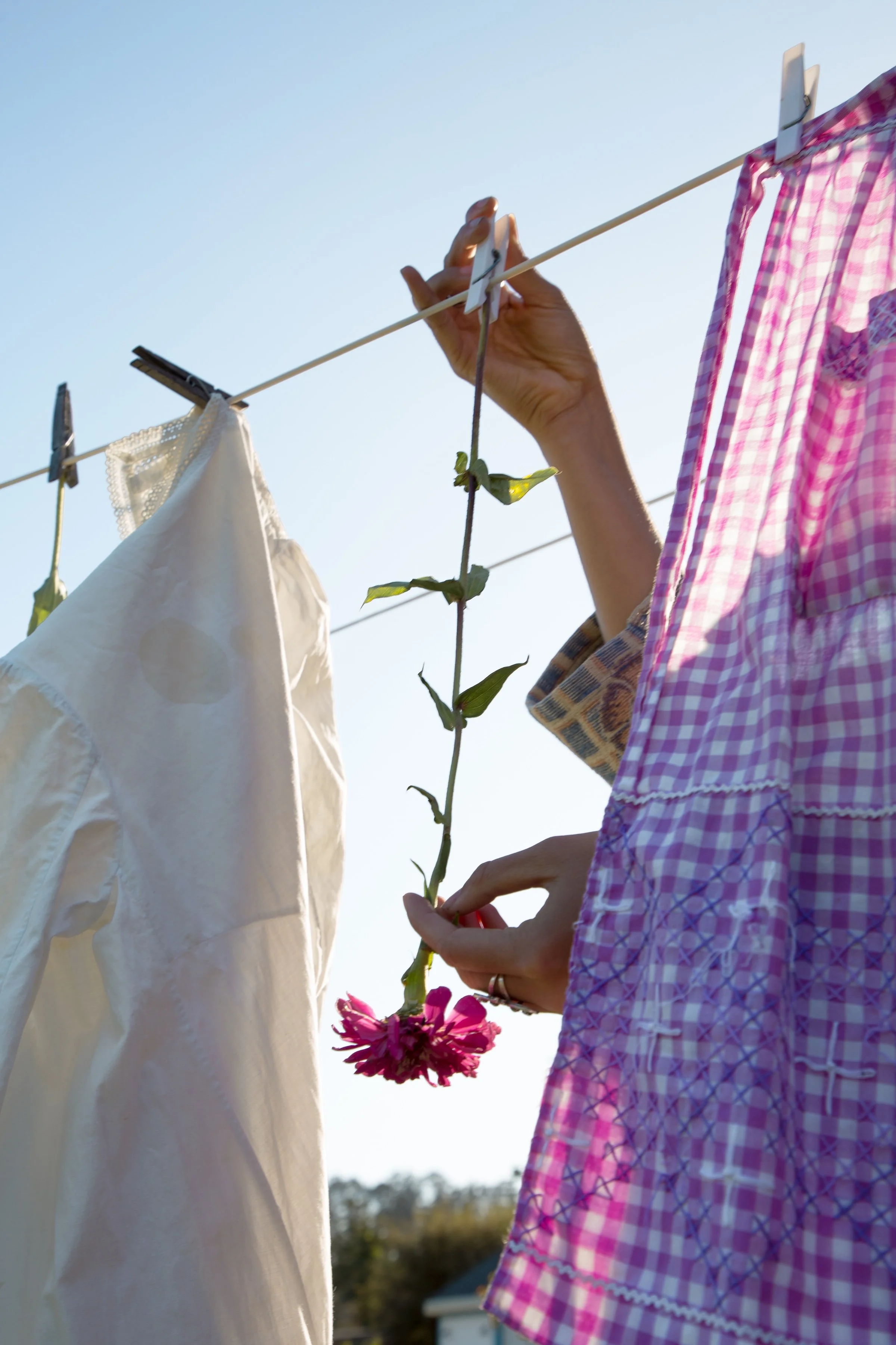 Person hanging pink flowers on a clothesline with clothespins, wearing a pink checkered dress, under a clear blue sky.