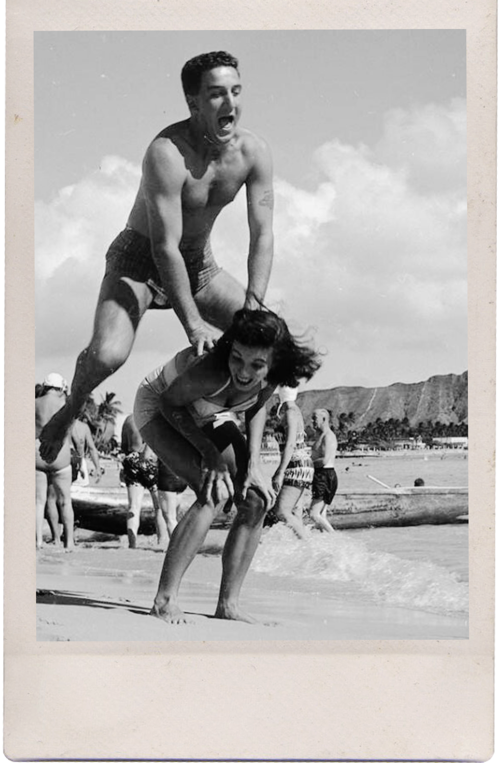 A black-and-white photo of a man and woman playing on a beach, the man standing on the woman's back and holding her hair while she bends forward, both smiling and laughing, with a mountainous coastline and several people in the background.