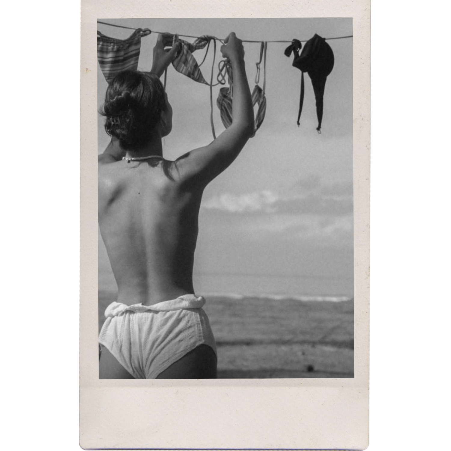 A black and white photograph of a man seen from behind, shirtless, hanging laundry on a clothesline with the ocean and sky in the background.