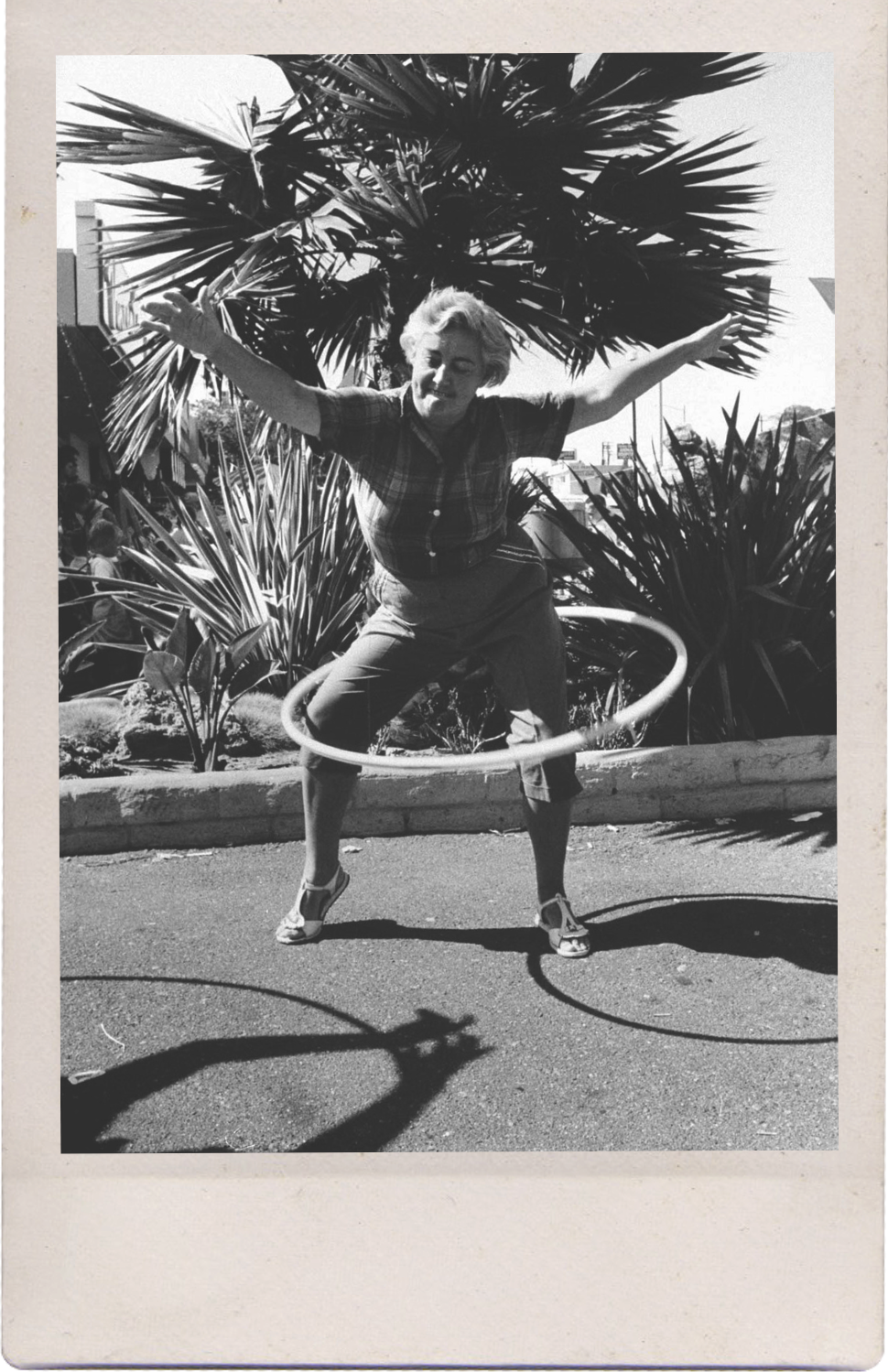 A woman smiling and playing with a hula hoop on a sidewalk with plants in the background.