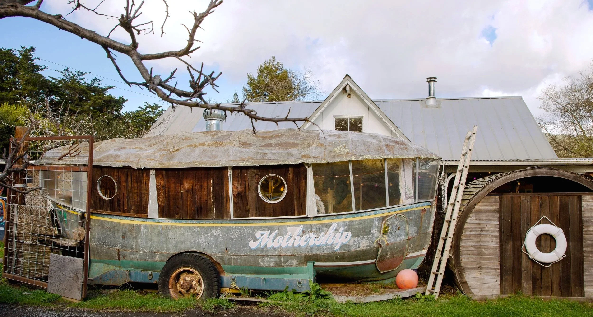 An old boat with the word "Mothership" painted on the side, situated outdoors near a wooden fence, with a house and trees in the background.