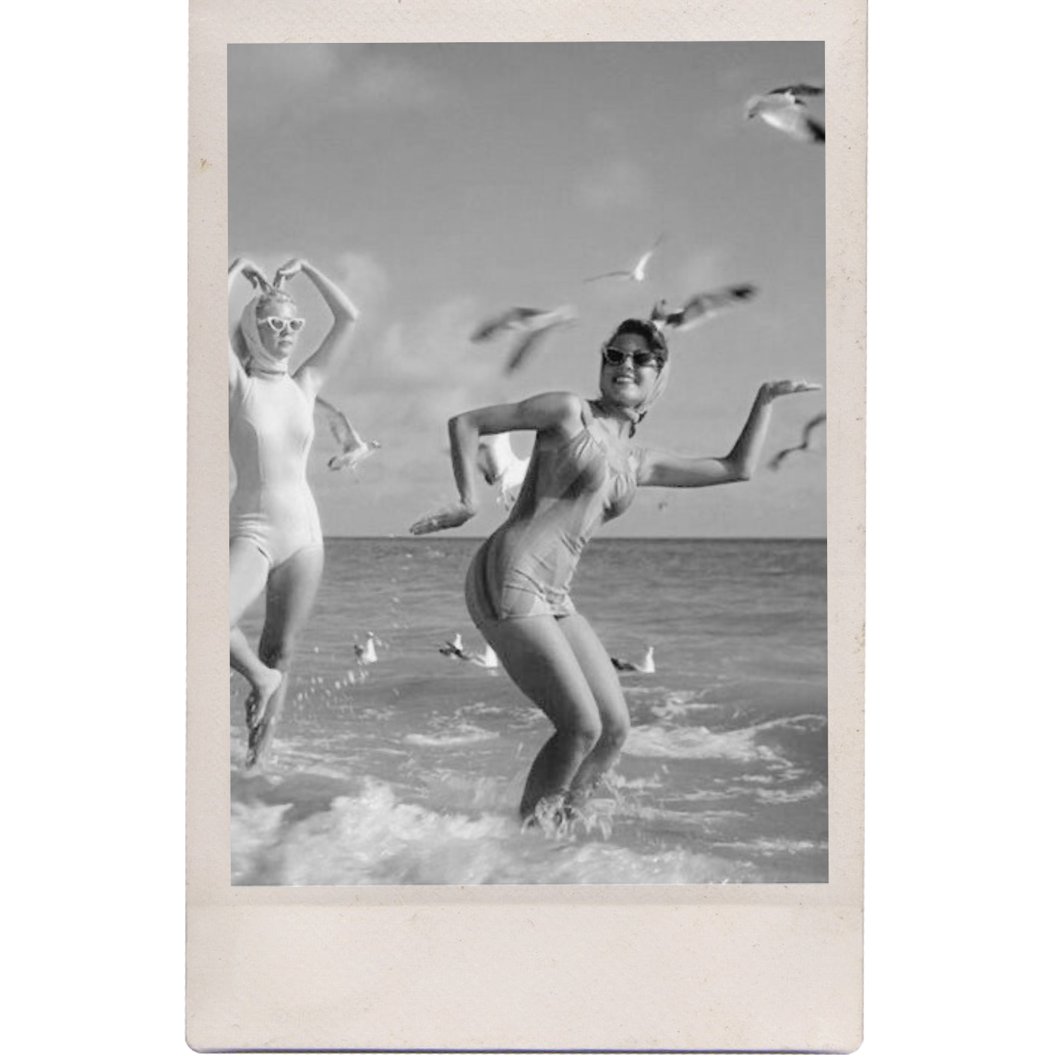 Black and white photo of two women in swimsuits running in the ocean, with seagulls flying overhead.