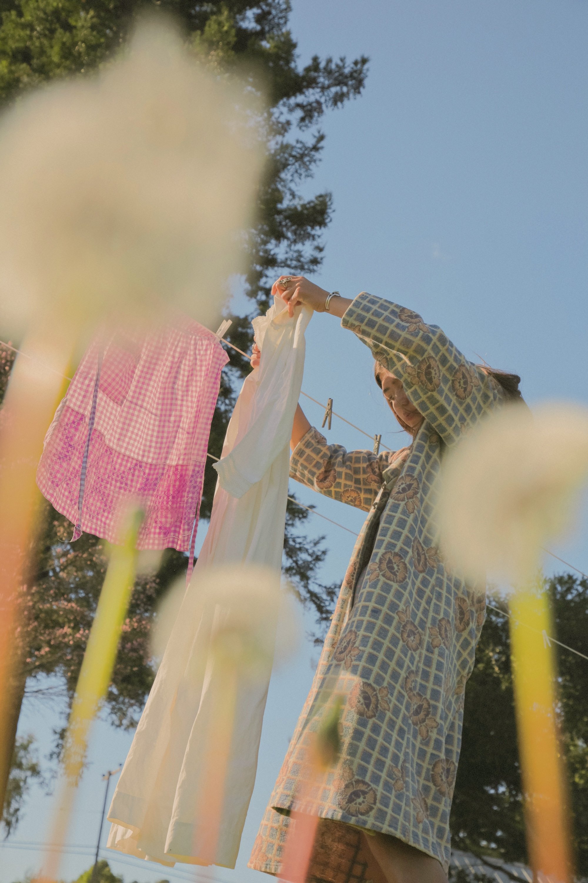 A woman hanging laundry on a clothesline outdoors against a clear blue sky, with trees in the background.