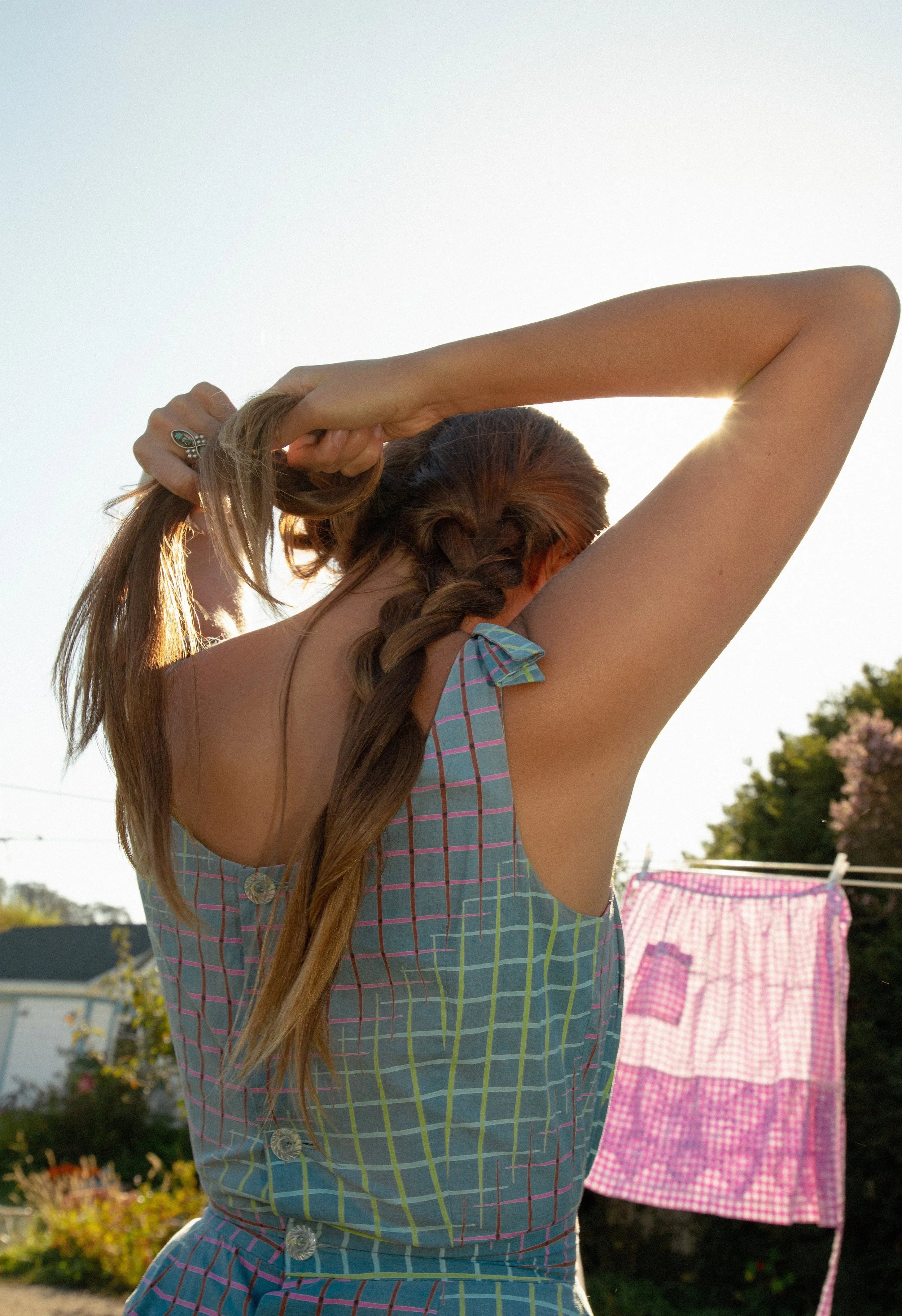 A woman with long hair tied in a braid, wearing a plaid dress, is outside holding her hair with both hands. The sun is shining behind her, and there are bushes and laundry with pink checkered patterns in the background.