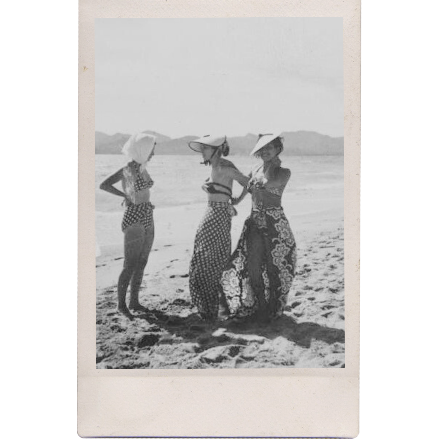 Four women wearing patterned skirts and head coverings stand on a sandy beach, facing each other and holding hands. They are under umbrellas with a mountain landscape in the background.
