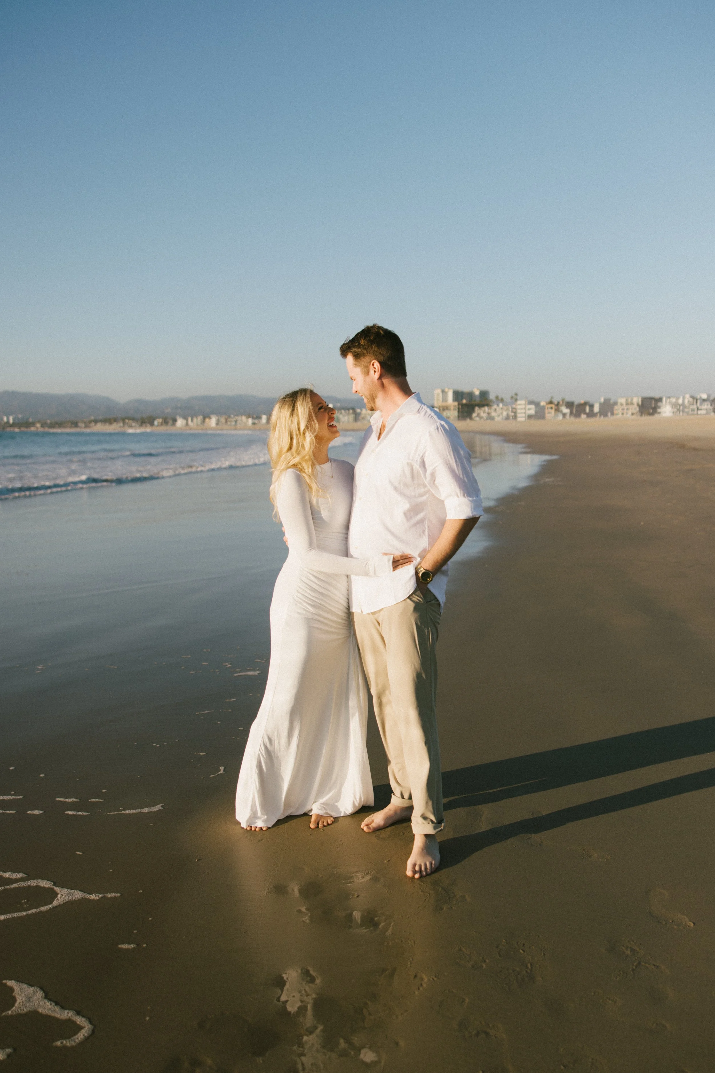 A happy couple in wedding attire standing barefoot on the beach, looking at each other lovingly, with the ocean and distant city skyline in the background during what appears to be late afternoon or sunset.