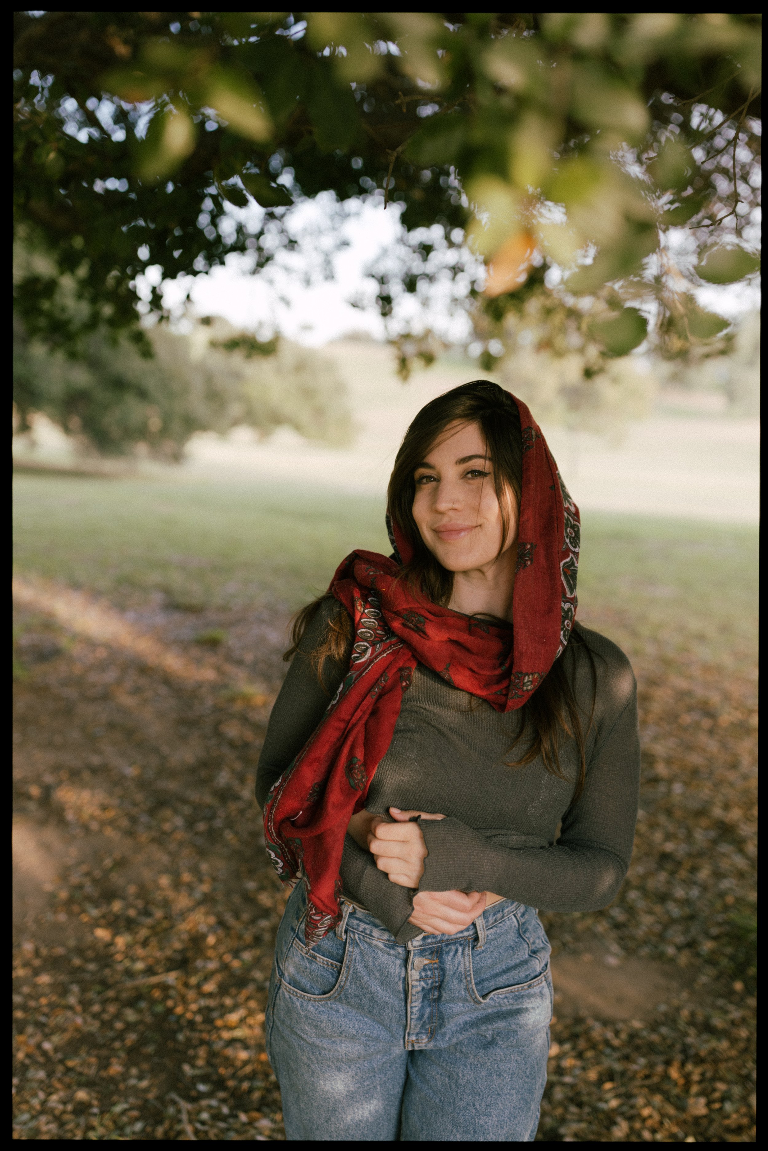 A young woman with dark hair and a scarf standing outdoors under a tree, smiling softly.
