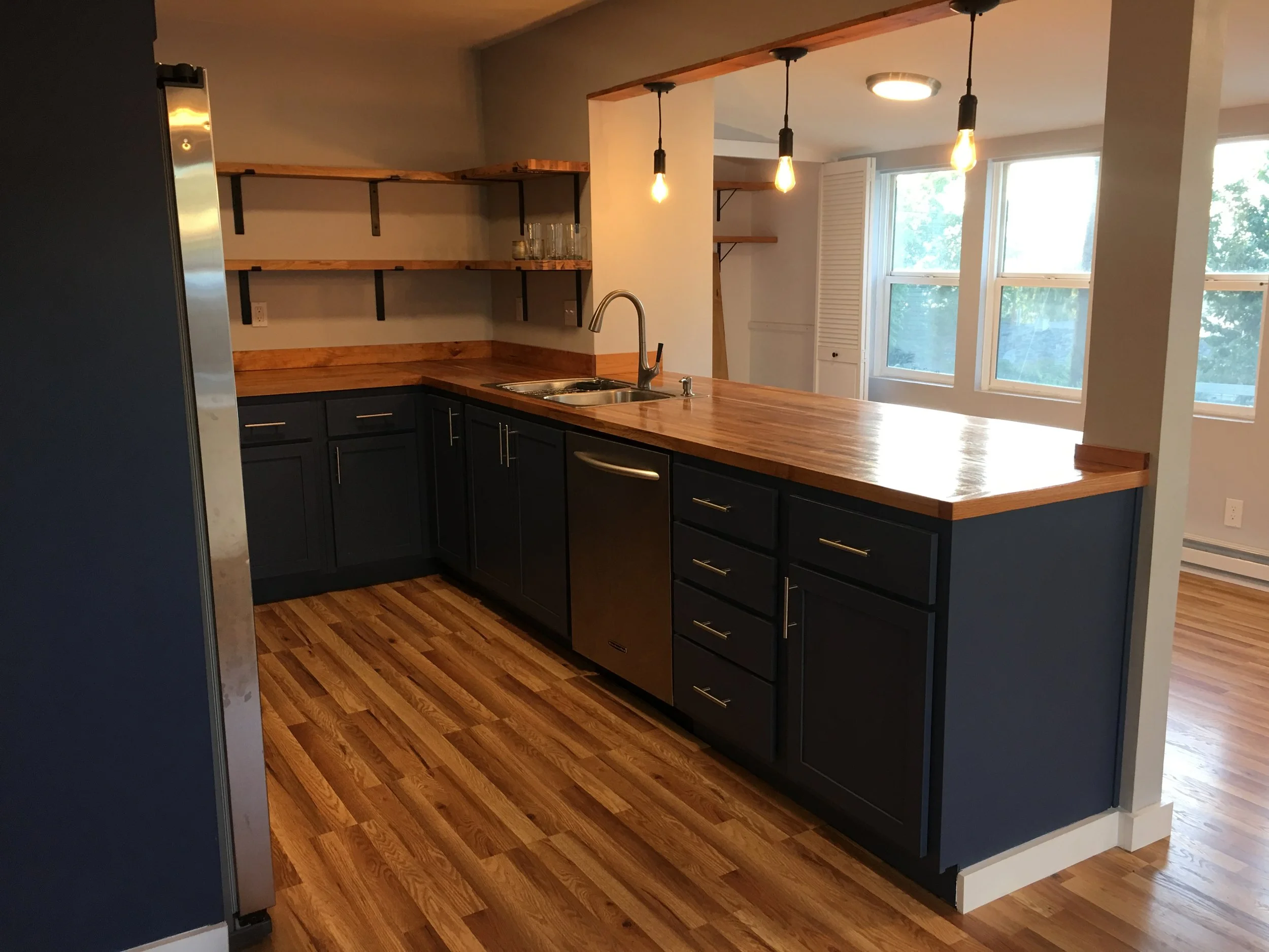 Modern kitchen with black cabinets, wooden countertops, open shelving, hardwood floors, and a large window letting in natural light.