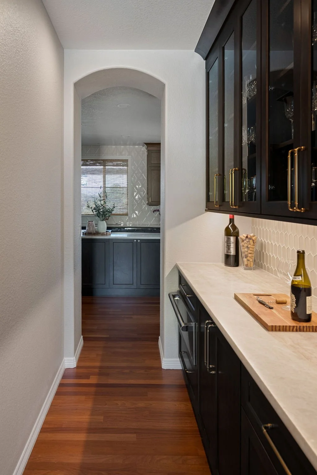 Elegant wine bar with dark woodland cabinetry, stone countertops, glass storage, and an arched entry to the kitchen.