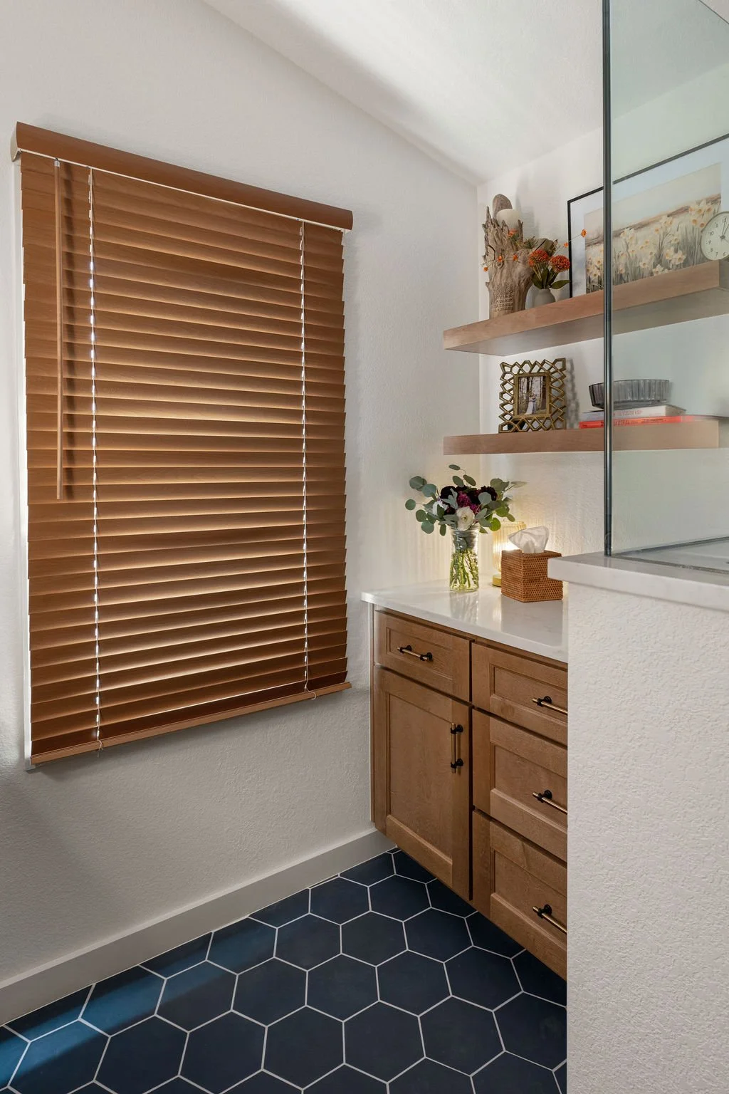 Remodeled bathroom with natural wood cabinetry, floating wood shelves, blue hexagon floor tile, and warm wood window blinds