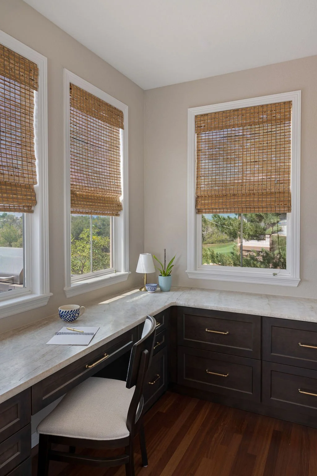 Bright home office nook with dark wood cabinetry, light countertops, large windows, and woven shades.