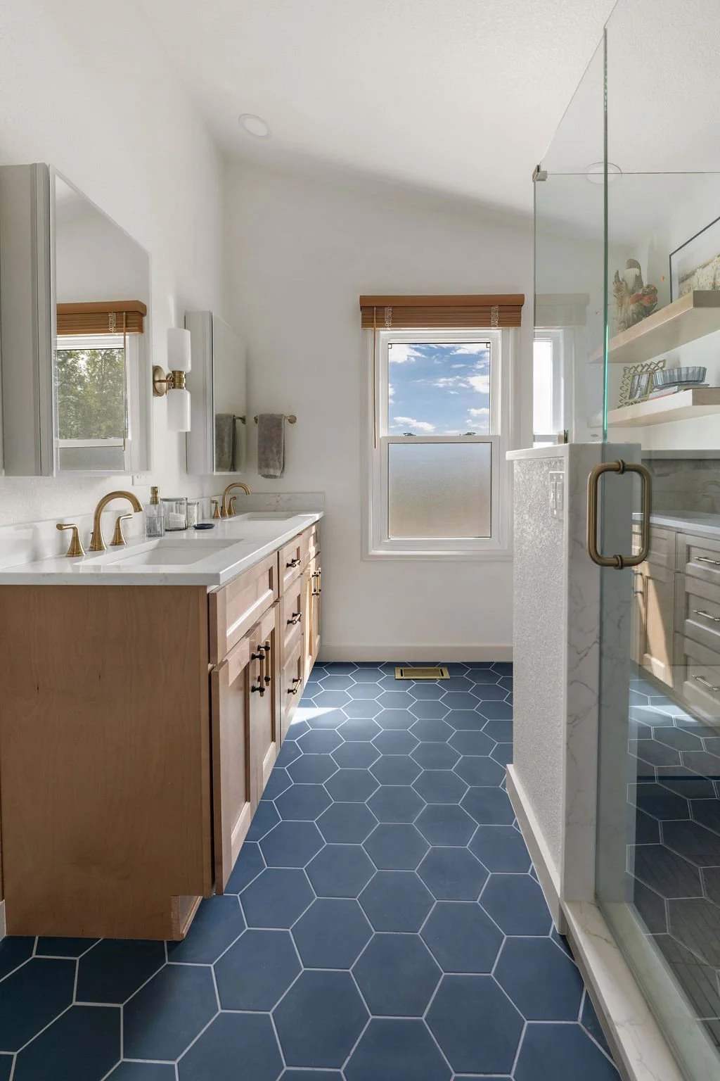 Remodeled bathroom with natural wood double vanity, glass walk-in shower, brass fixtures, and blue hexagon floor tile