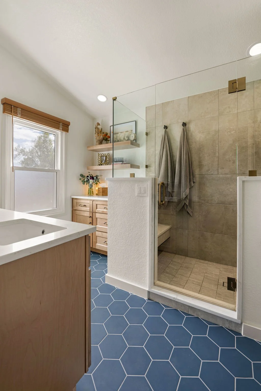Remodeled bathroom with walk-in glass shower, natural wood vanity, floating shelves, and blue hexagon floor tile