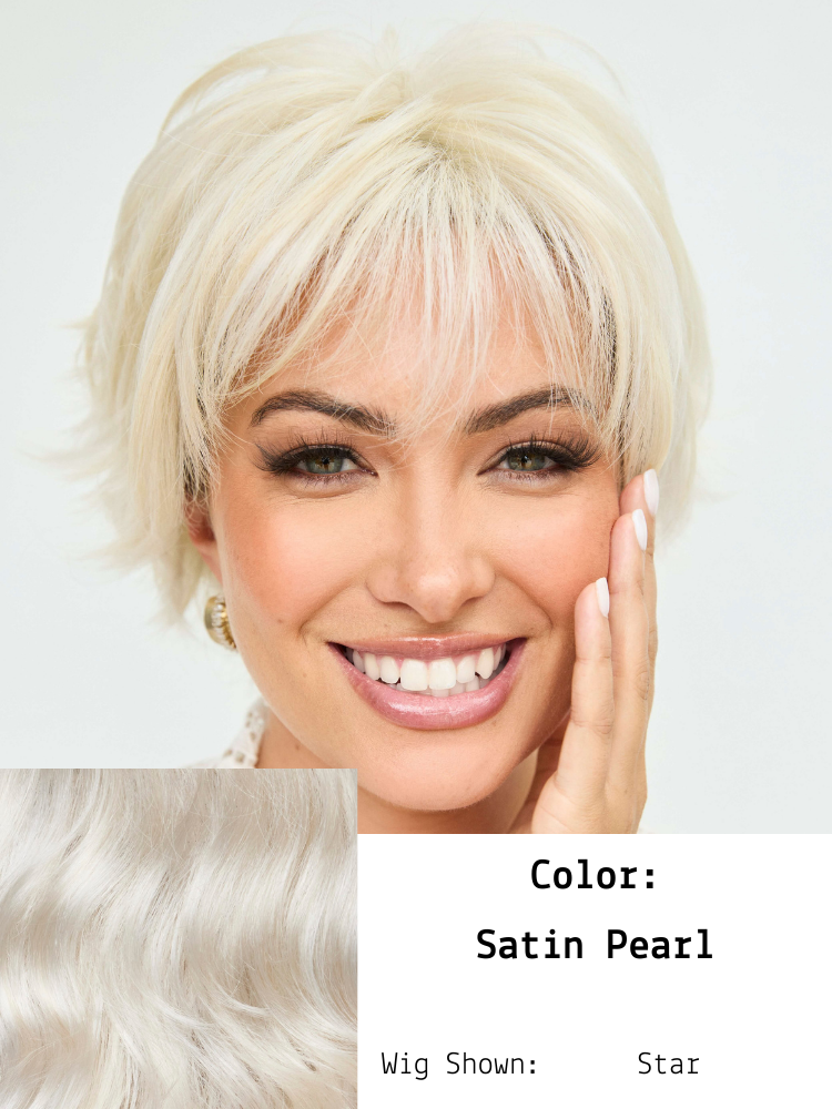 A woman with short, platinum blonde hair styled in a layered pixie cut, smiling with her hand gently touching her face, wearing makeup with defined eyebrows and lipstick. An inset shows her hair wig in satin pearl color with soft waves. The text indi