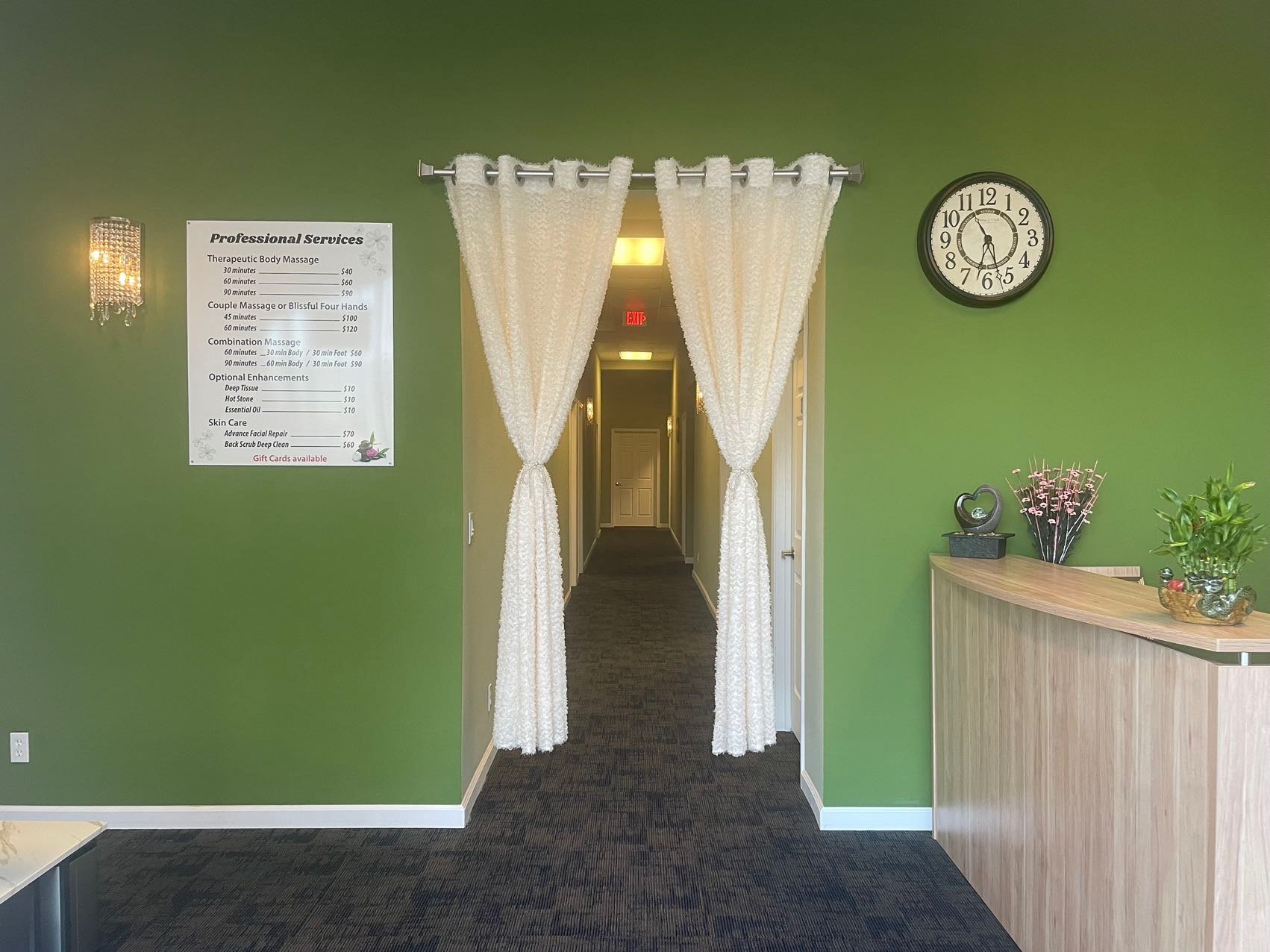 Interior of a massage spa with green walls, a list of services, a clock showing 12:15, a reception desk with plants, and a hallway with white curtains.