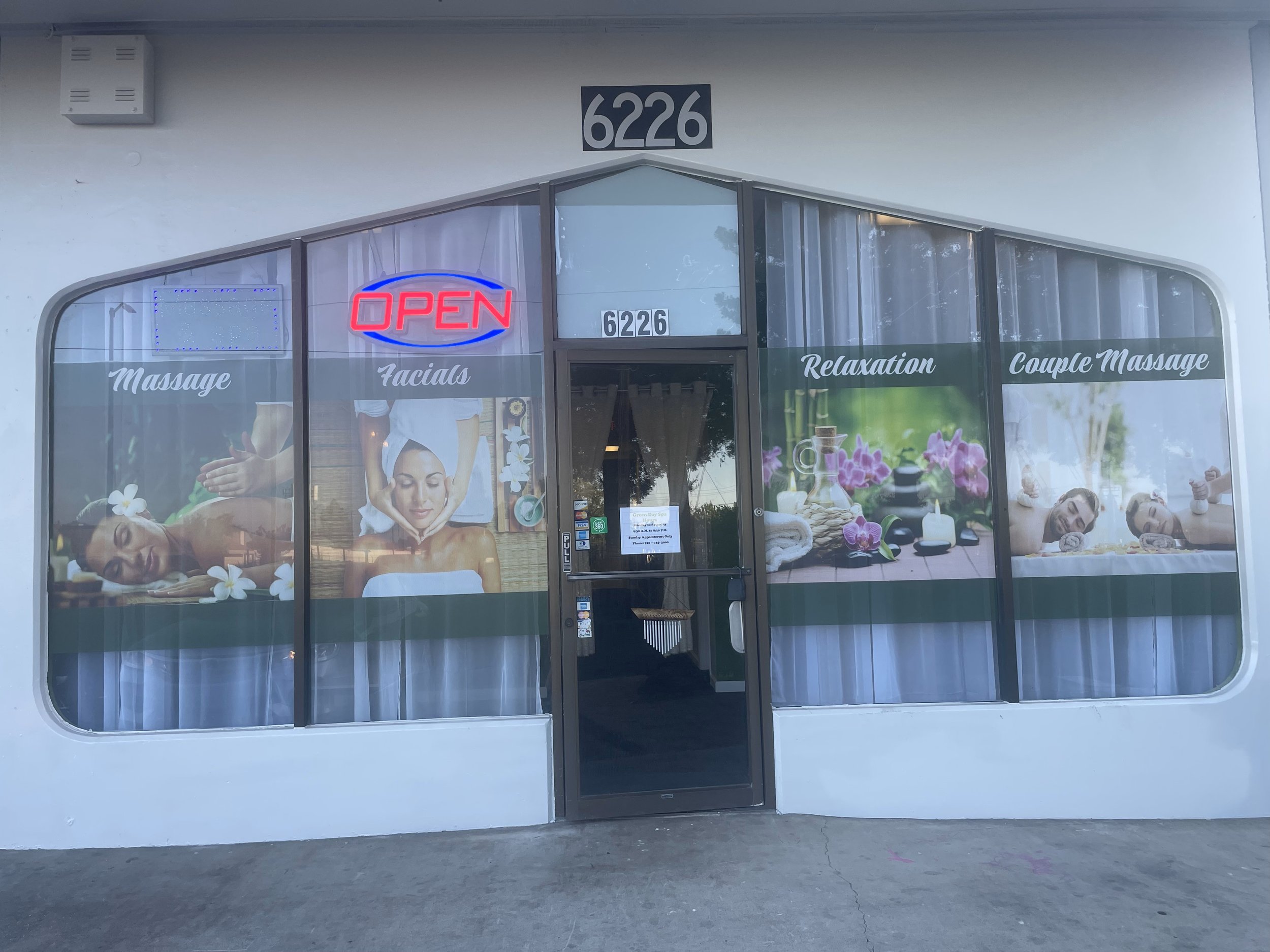 Front of a spa with window advertisements for massage and facial services, displaying images of people receiving massages. The door has a sign with the spa's address, 6226, and a neon "OPEN" sign is lit.