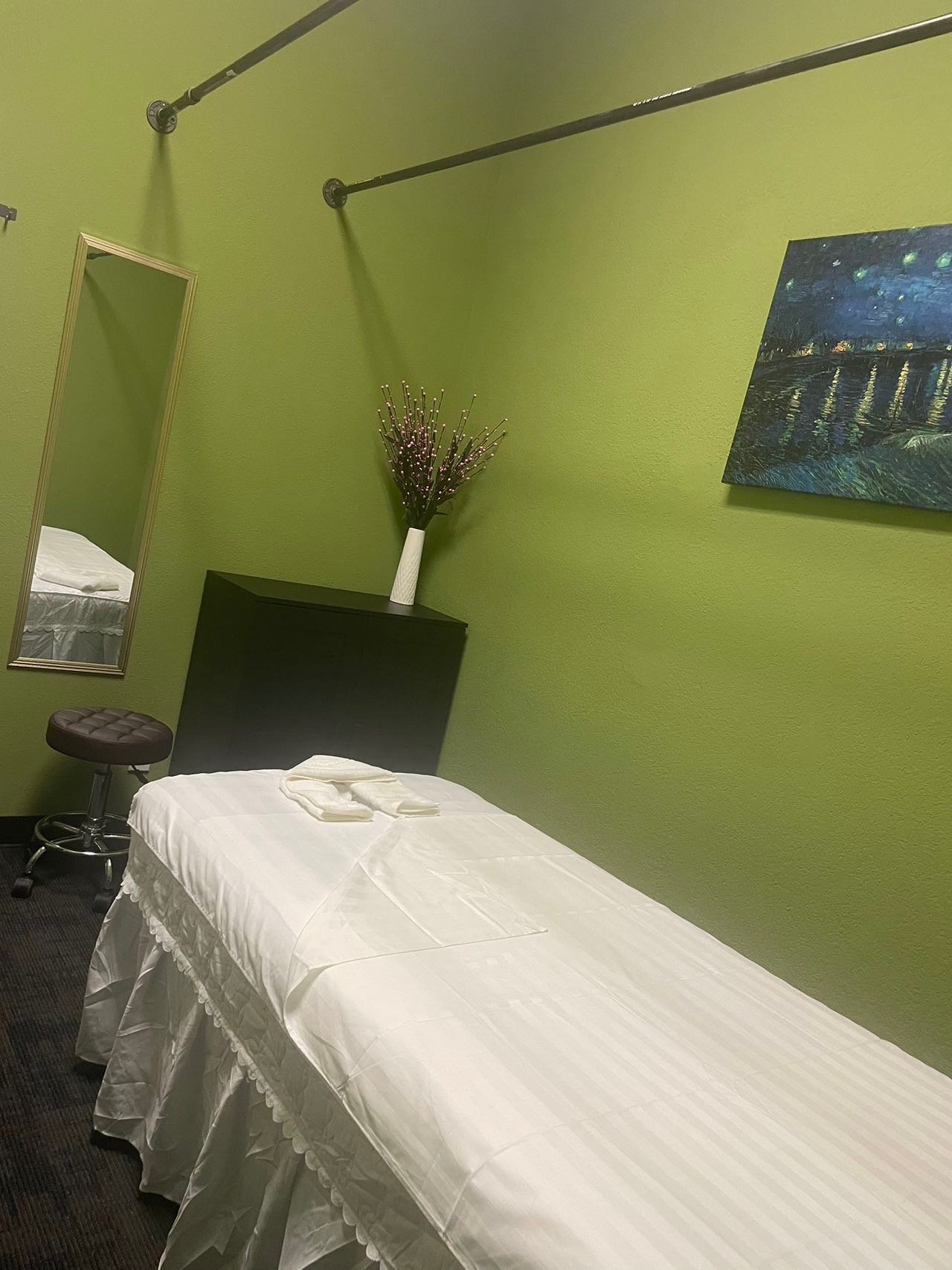 Massage room with a green wall, a massage table covered with a white sheet, a stool, a framed mirror, a painting on the wall, and a vase with decorative branches.