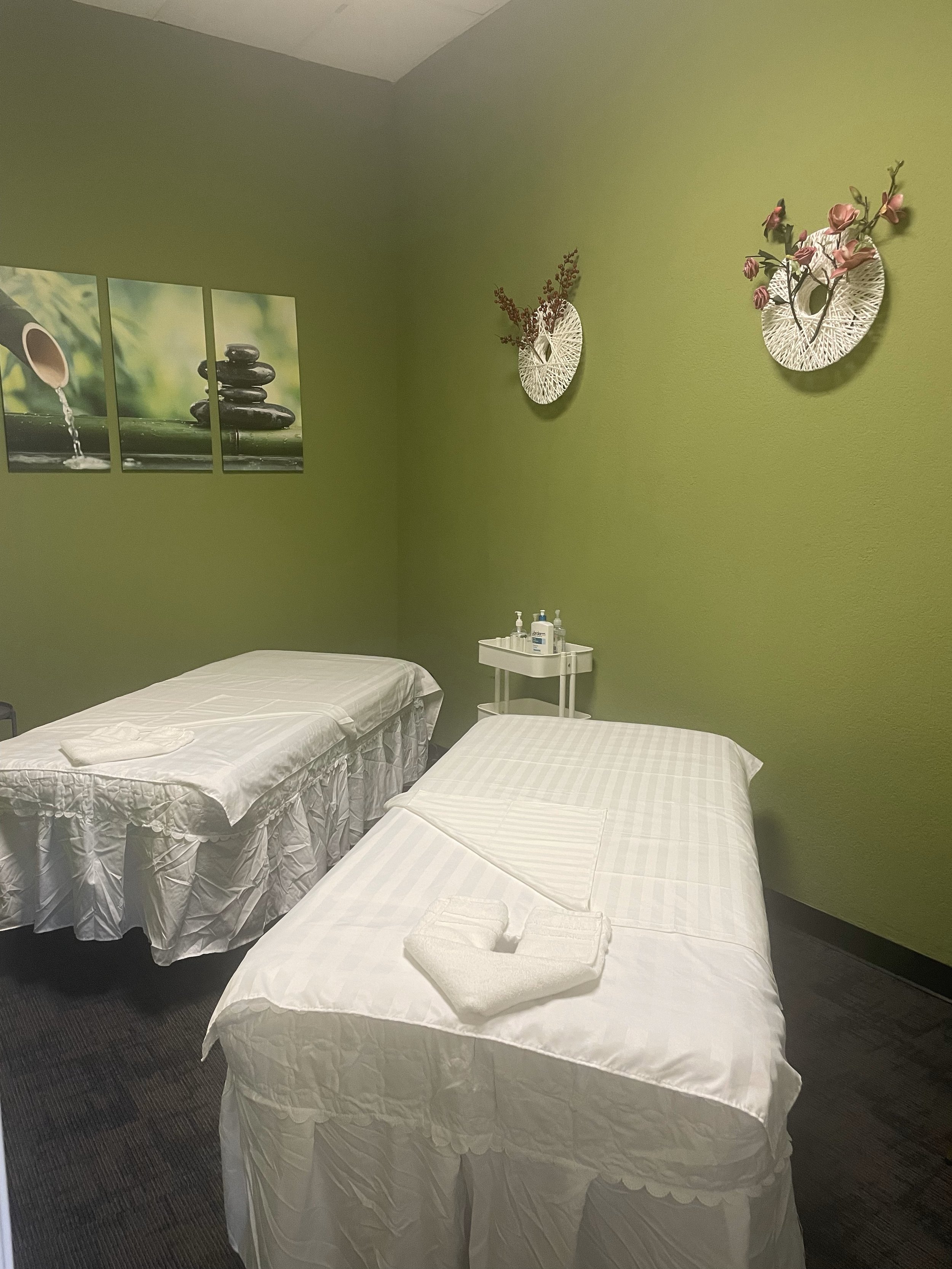 Massage therapy room with two beds, white linens, and a green wall featuring relaxing artwork. Decor includes wall-mounted floral arrangements.