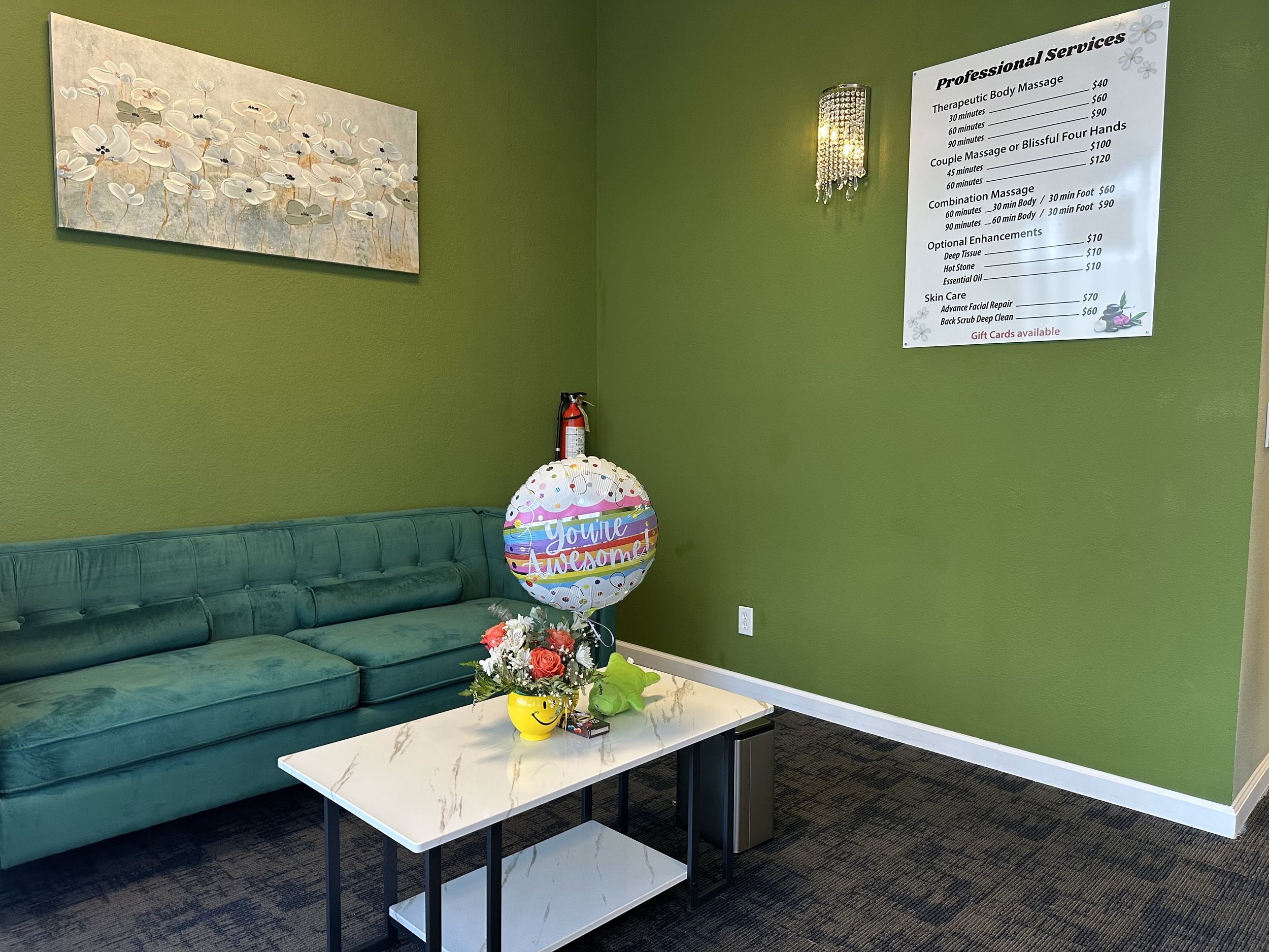 Interior of a waiting area with a green sofa, marble coffee table, and decorative balloon. A flower painting is on a green wall, alongside a menu of professional services and a crystal wall light.