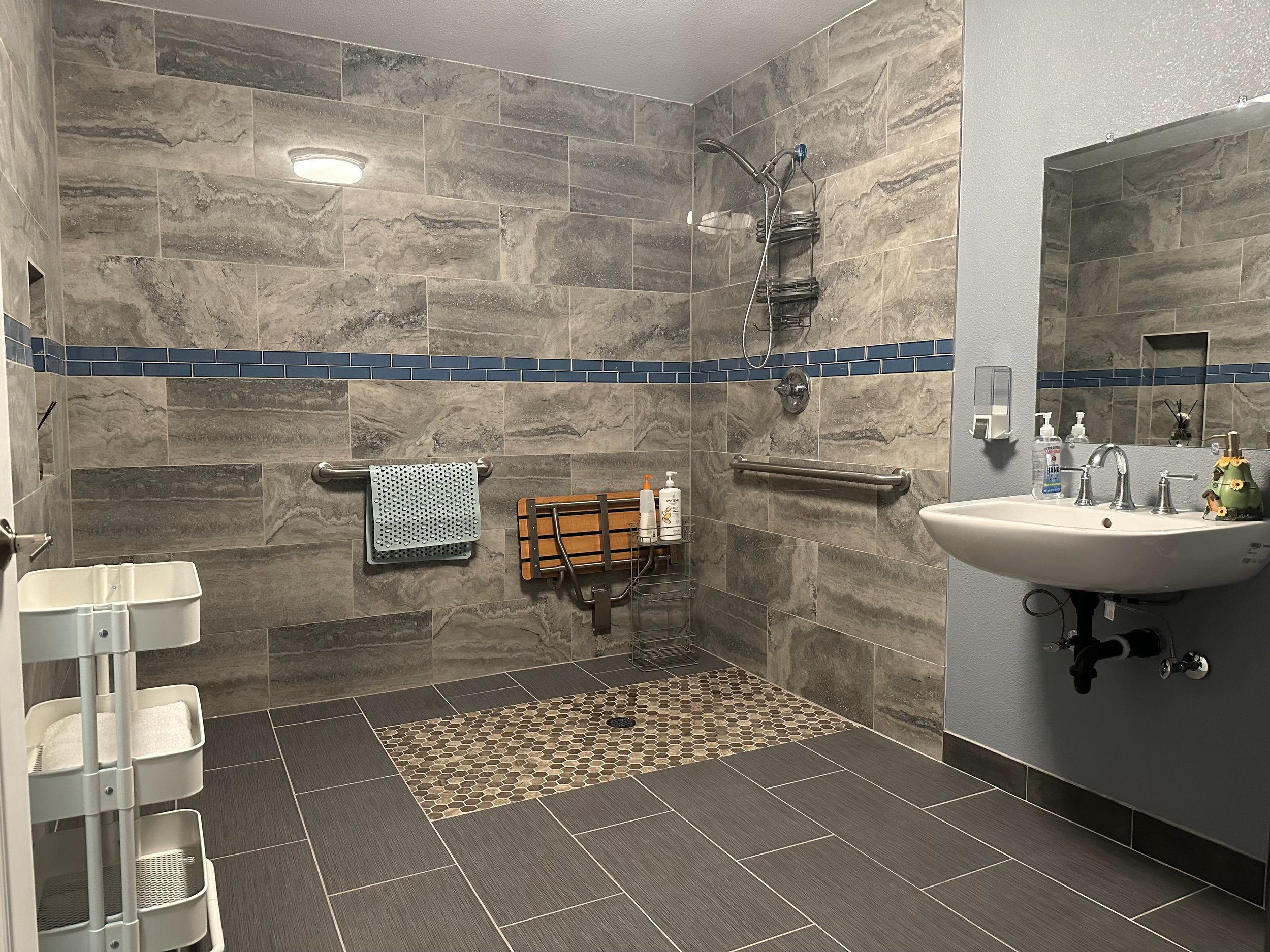 Modern accessible bathroom with gray tile walls, a walk-in shower, a wall-mounted bench, handrails, and a sink on the right. Hygiene products are visible on a shelf, and a towel is hanging from the handrail.