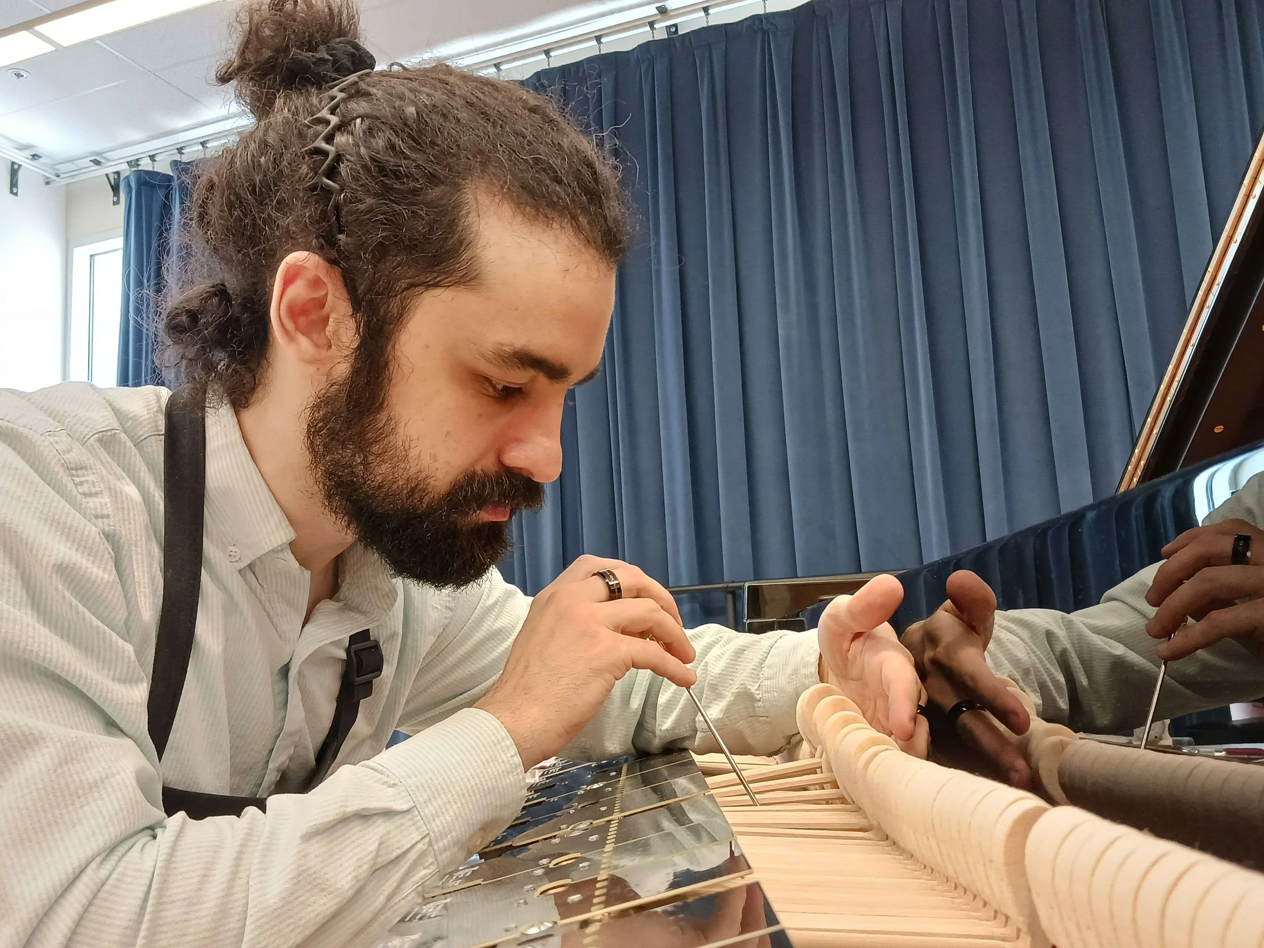 A man with long, curly hair, a beard, and a mustache, wearing a white striped shirt and black suspenders, is working on the inside of a piano. He is using a tool to work on the piano strings or interior components, with a blue curtain backdrop.
