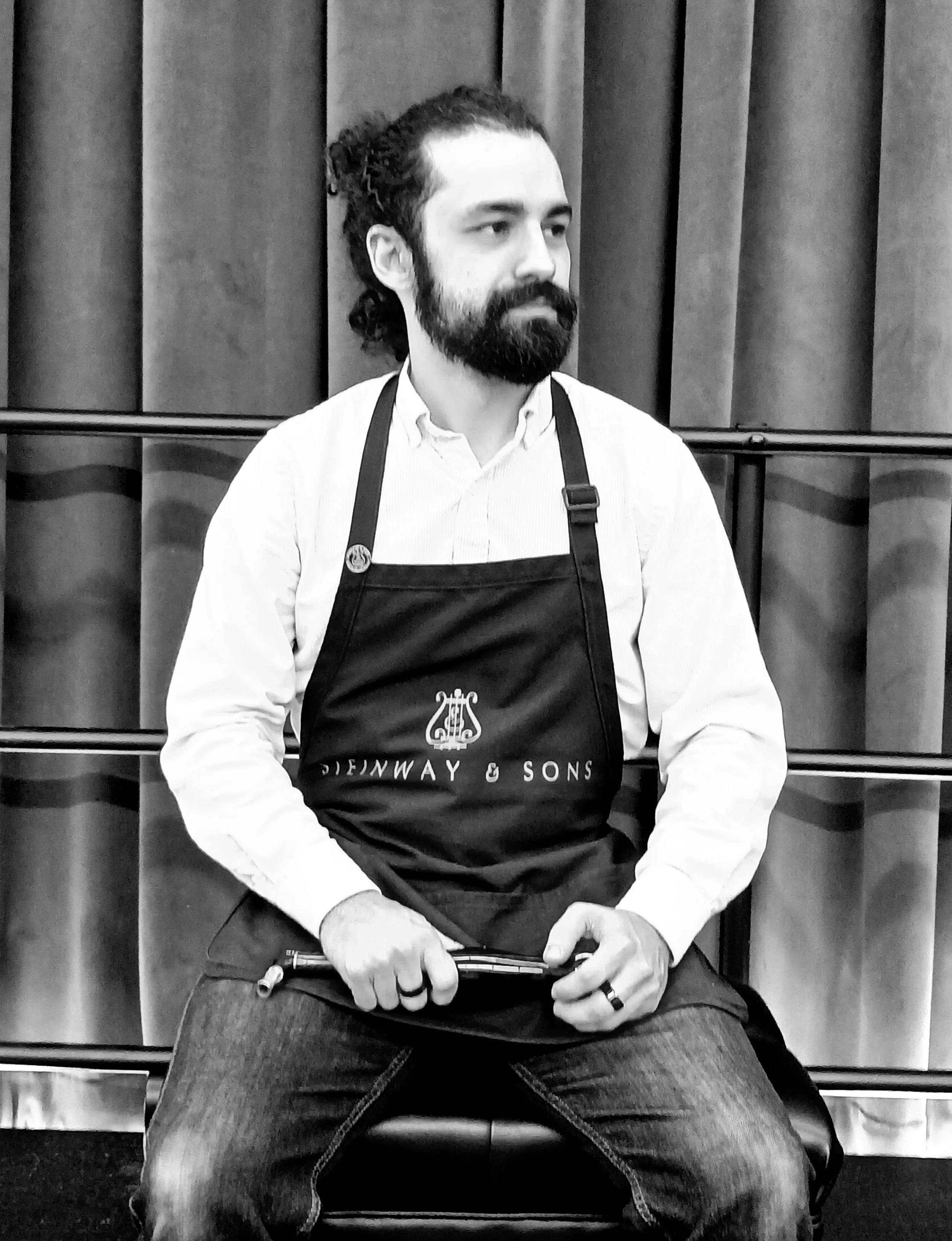 A man with a beard and long hair tied back, wearing a light-colored shirt and an apron with 'Steinway & Sons' logo, sitting indoors in front of a curtain or backdrop, holding a piano tuning lever.
