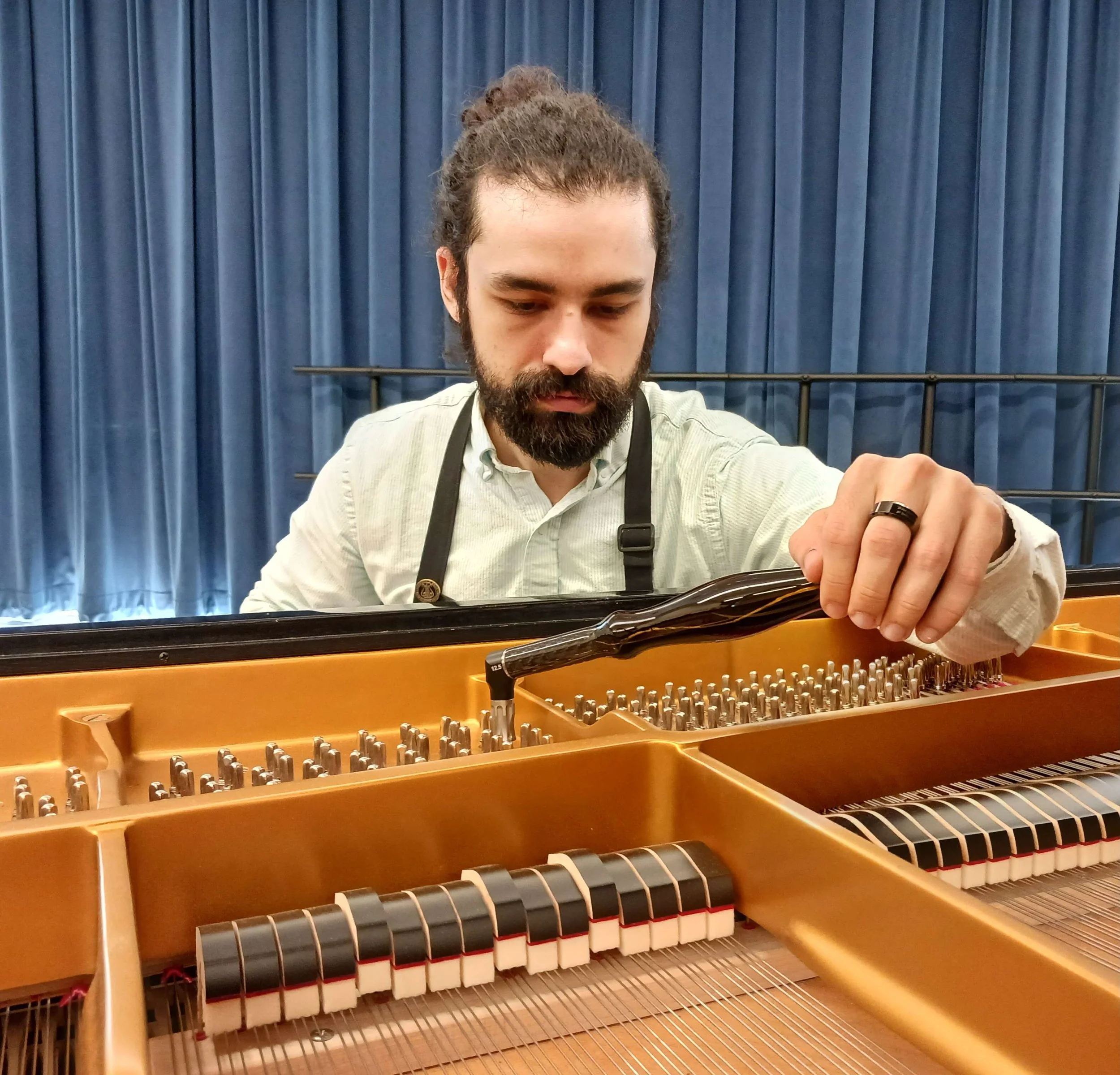 A man with a beard and long hair tied back, wearing a light-colored shirt and a ring on his hand, tuning the strings of a grand piano with a tuning hammer, in front of blue curtains.