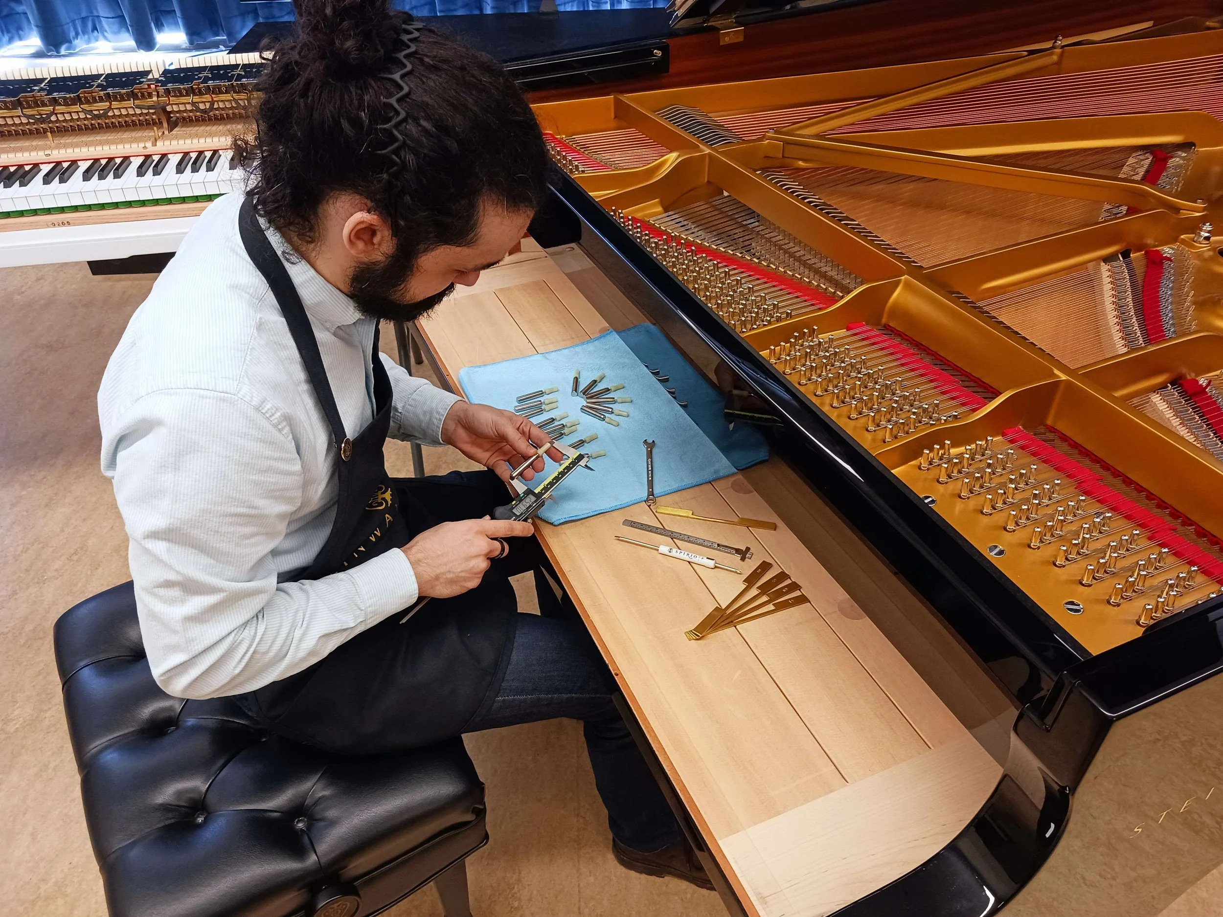A man sitting on a black stool working on a grand piano, with piano parts and tools laid out on the wooden work surface.