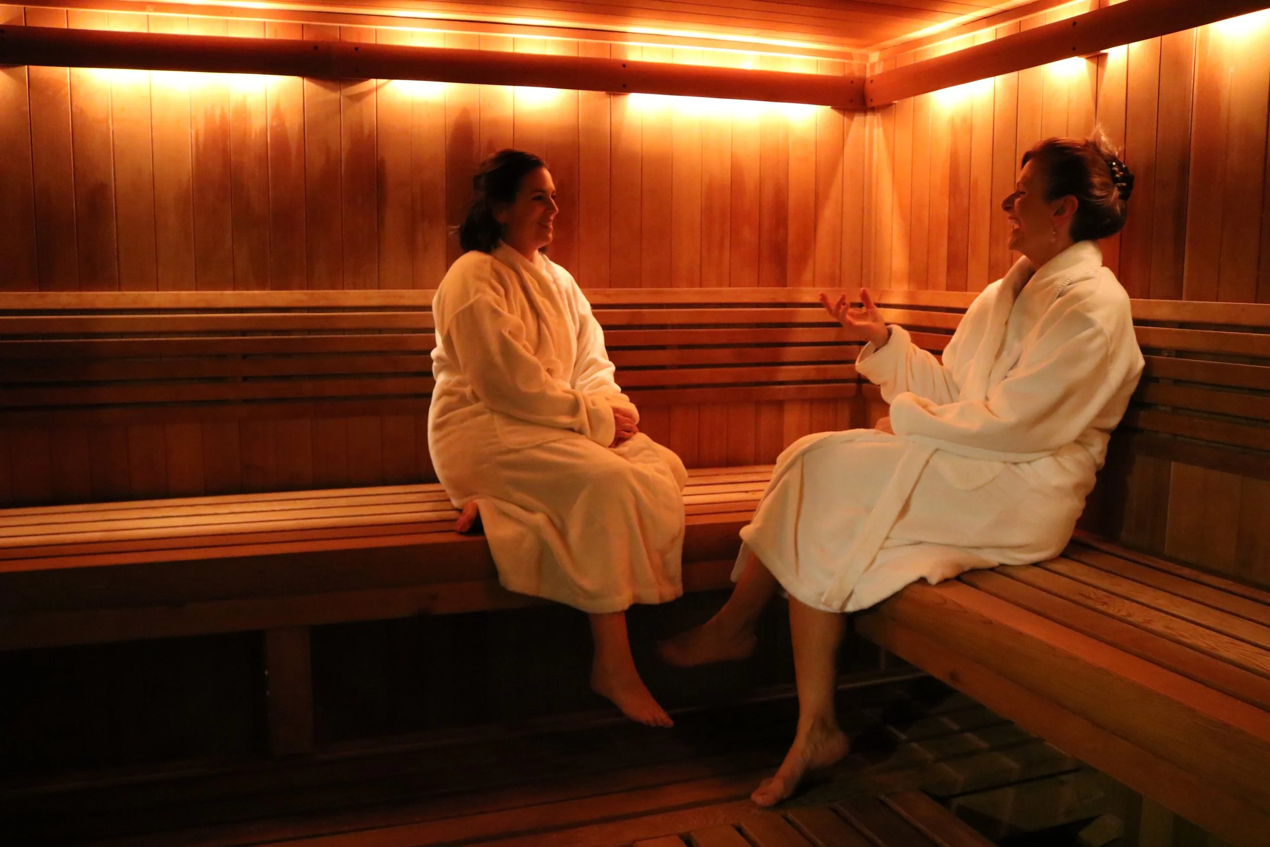 Two women sitting in a wooden sauna, wearing white robes, engaged in conversation and smiling at each other.