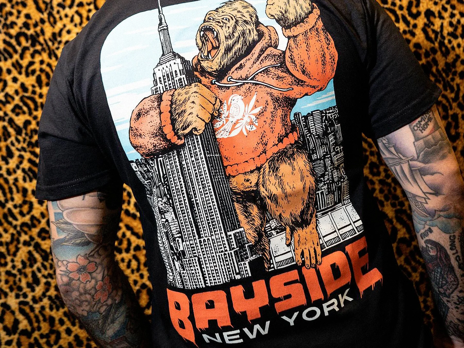 A person wearing a black T-shirt featuring a graphic of a muscular lion with a mane, holding the Empire State Building in one paw and a pistol in the other, with a cityscape background and the words 'ROCK SHOP' and 'NEW YORK' at the bottom, and tattoos visible on their arms.