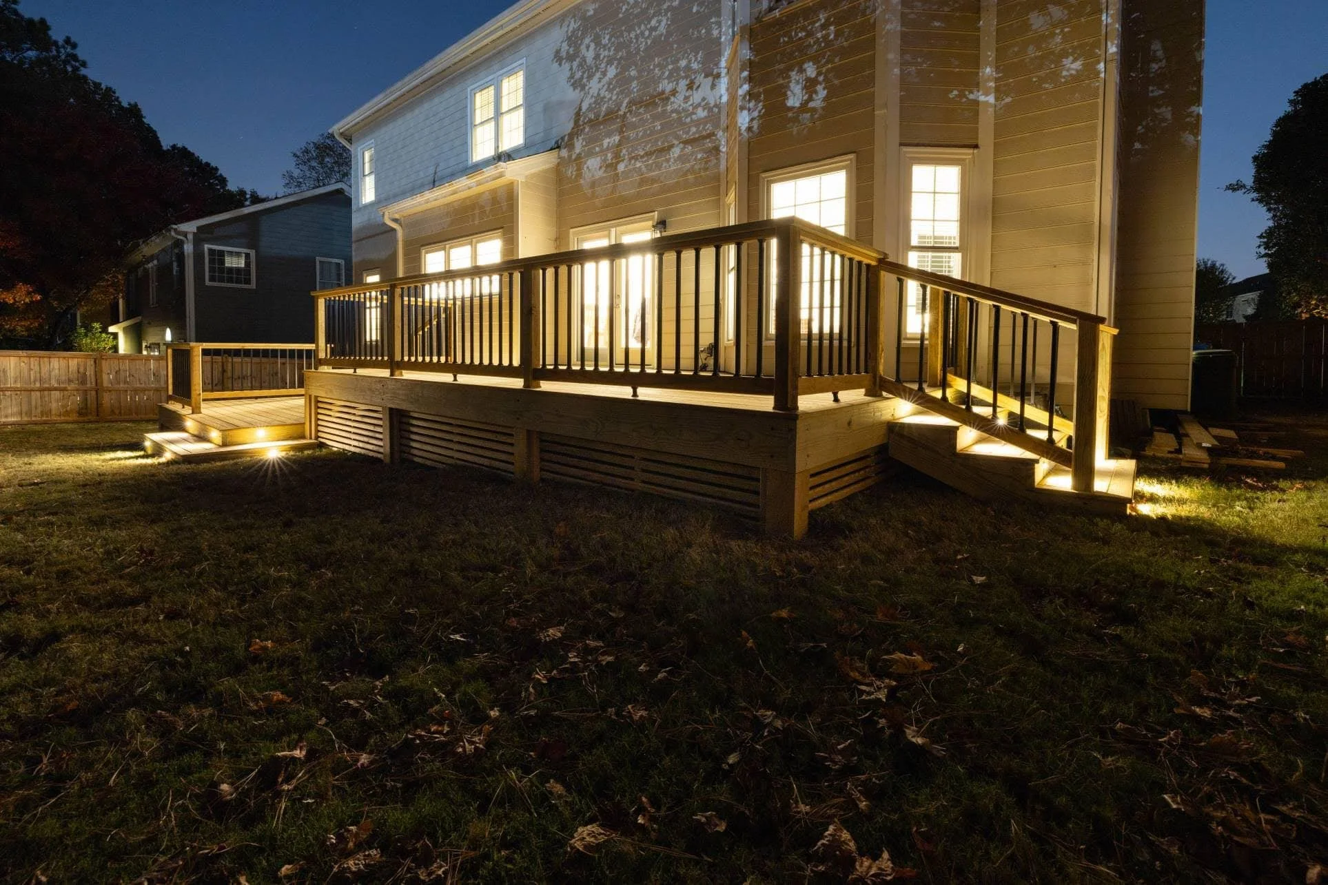 A wooden deck with lighting, attached to the back of a house, illuminated at night with a fence and neighboring houses visible.