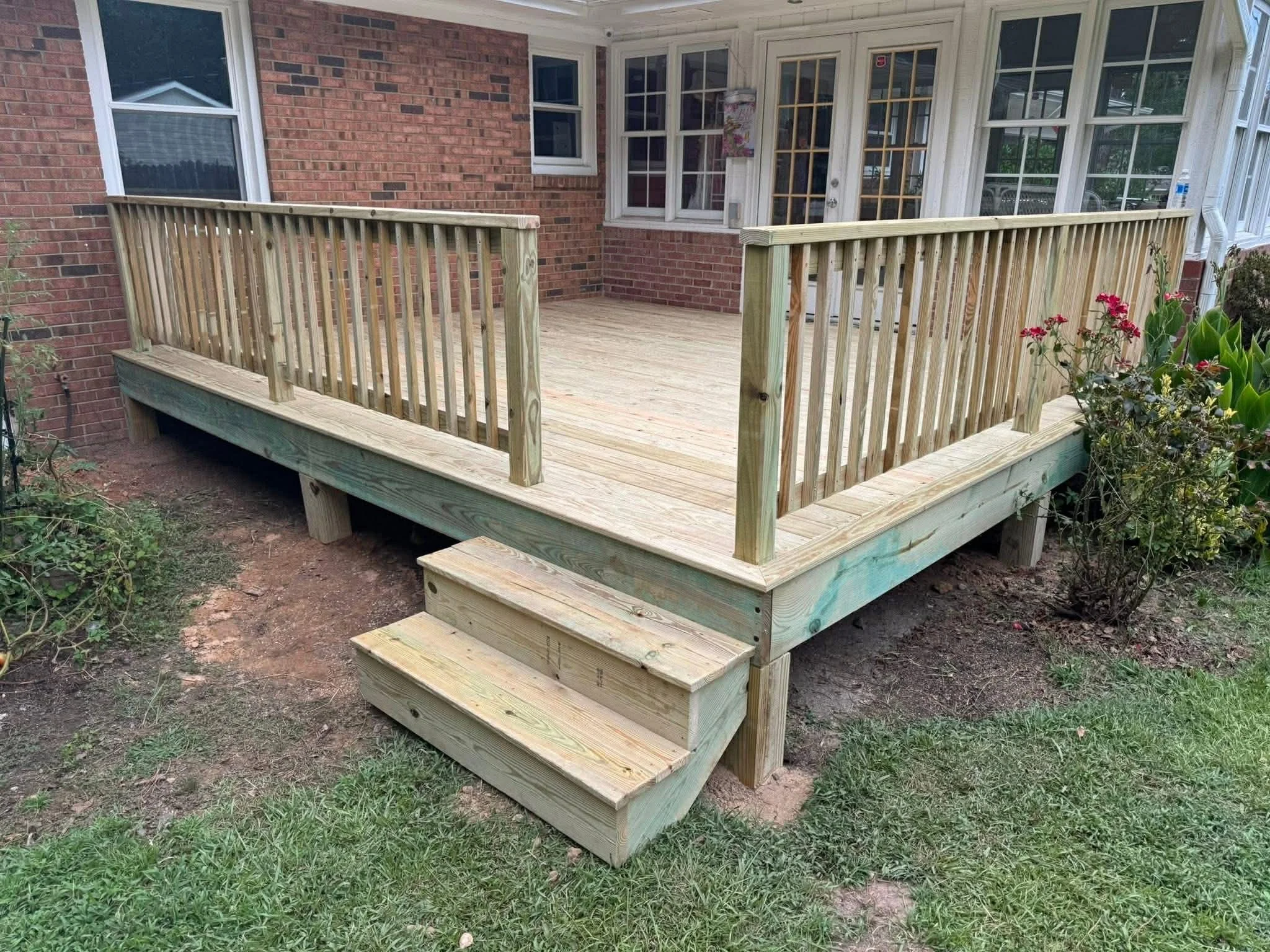 Newly built wooden deck with railings and steps outside a brick house, surrounded by plants and grass.