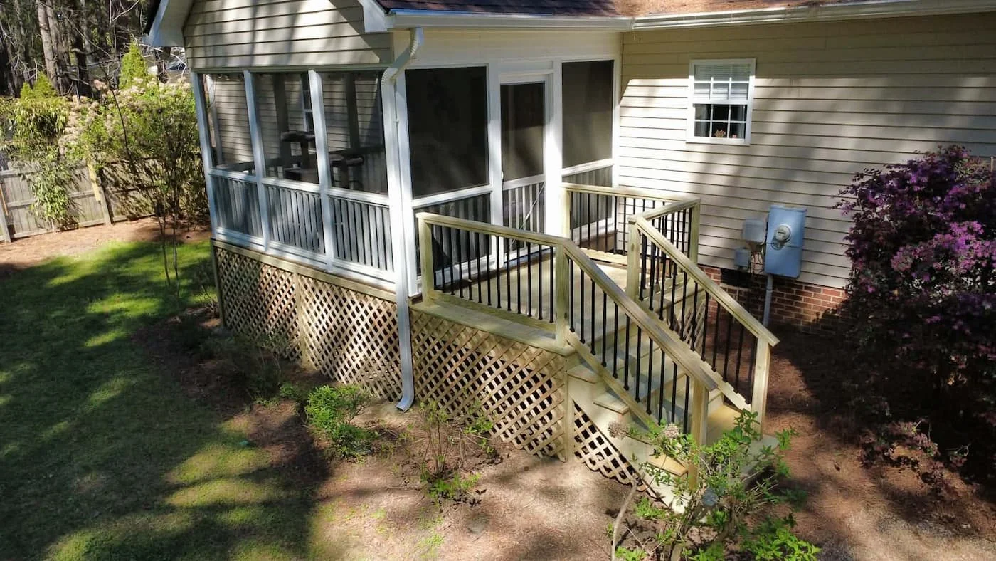 Newly built wooden staircase with black railing leading up to a screened porch on the back of a house with beige siding and a small window with flower decorations, surrounded by a landscaped yard with bushes and a flowering shrub.