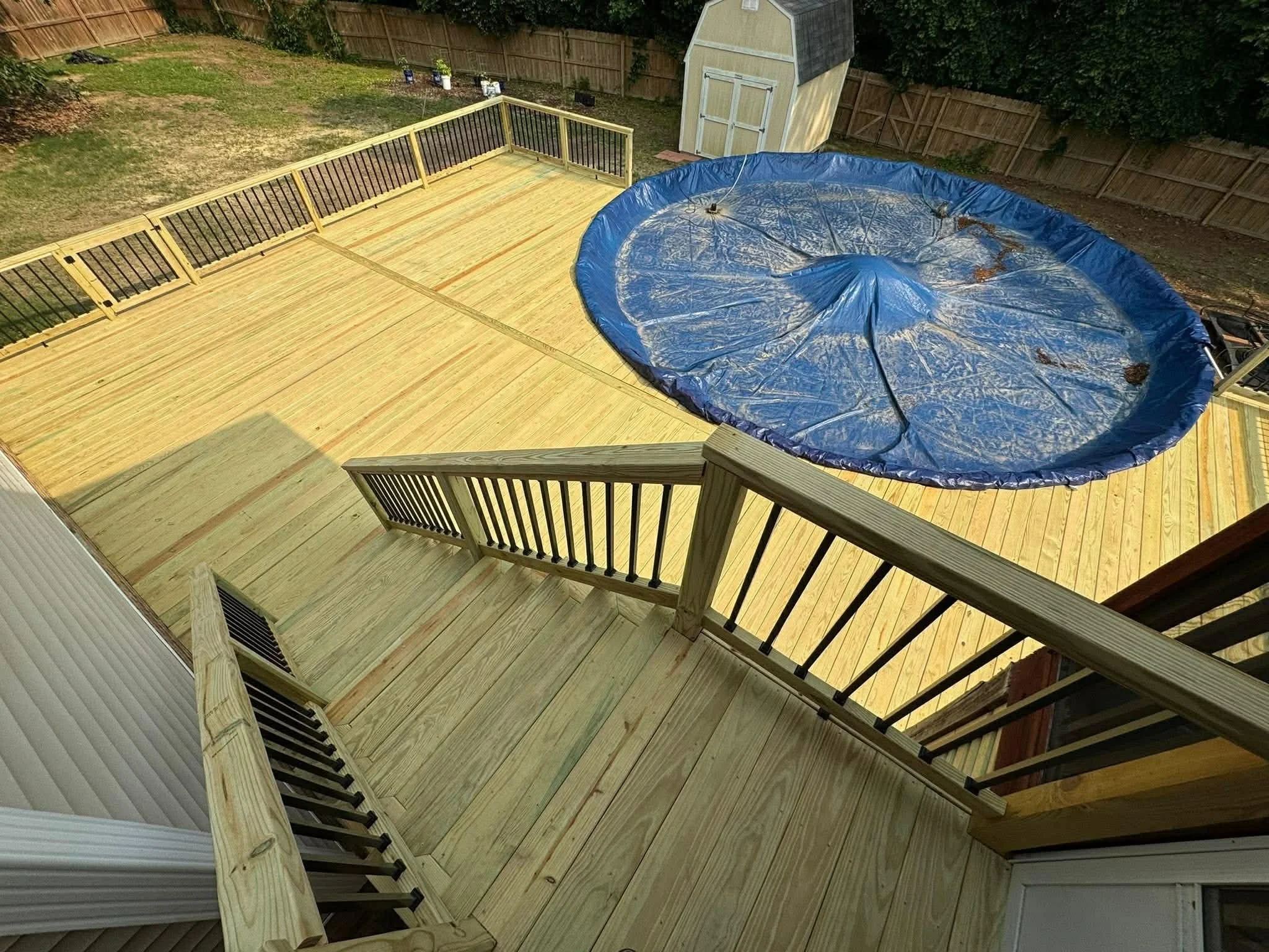 A wooden backyard deck with stairs, a fenced yard, a small shed, and a partially collapsed blue above-ground pool.