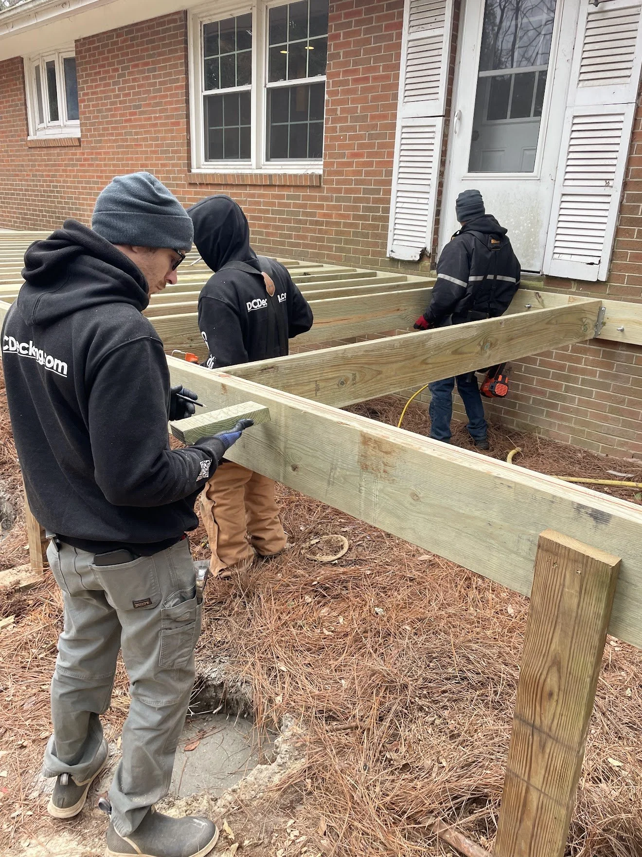 Four men constructing a wooden deck outside a brick house, wearing cold weather clothing and working with tools.