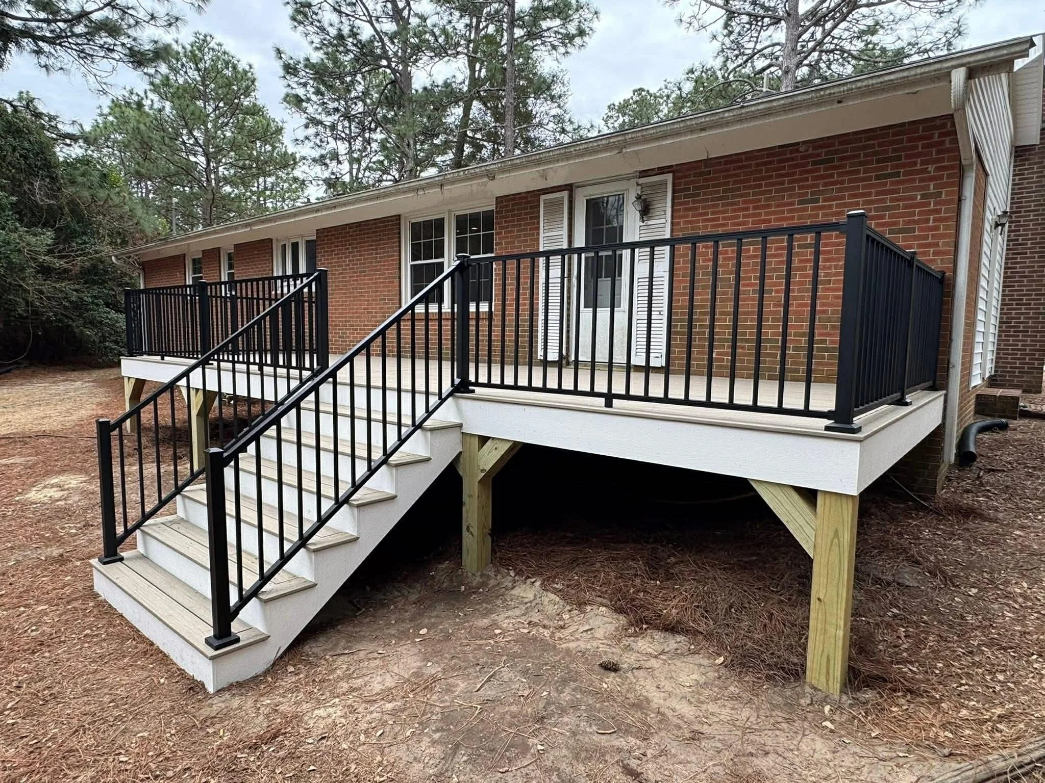 A newly built wooden deck with black metal railings and steps, attached to a brick house, surrounded by a wooded area with pine trees.