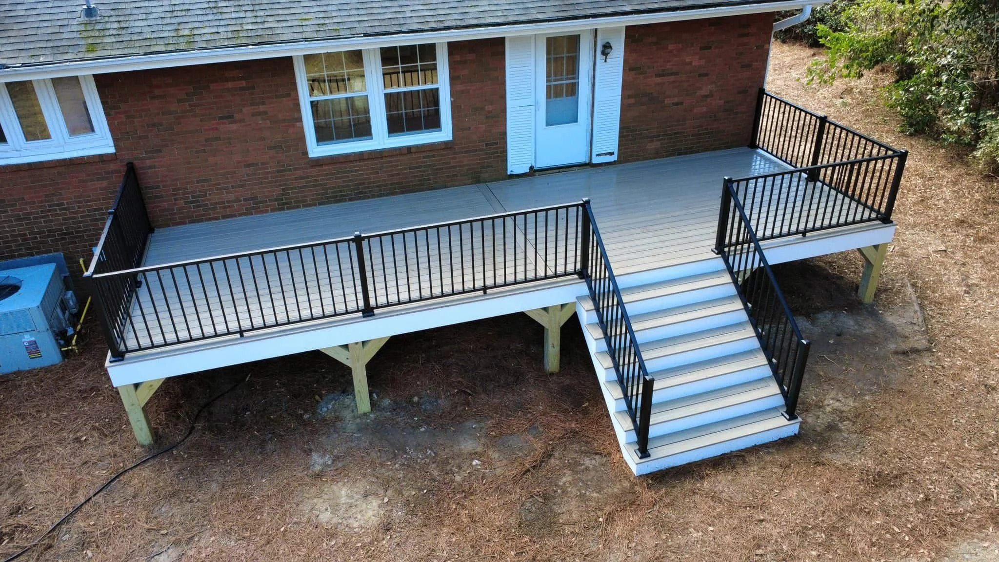 Newly built wooden deck with black railing around the perimeter, some leaves scattered on the surface, and steep stairs leading down from the deck. A trampoline is visible in the background, along with trees and neighboring structures.
