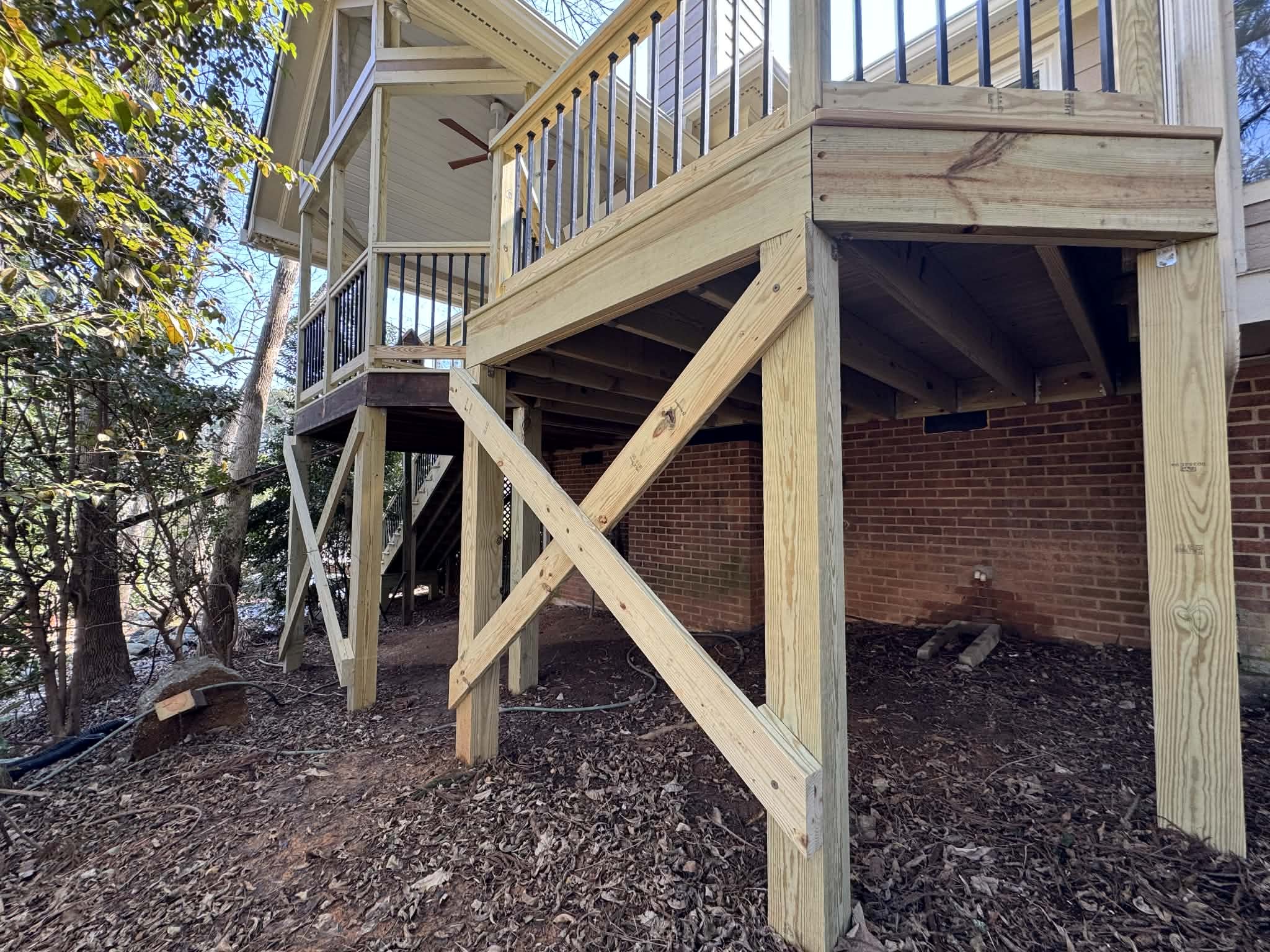 Newly constructed wooden deck with staircase under construction attached to a house with brick wall, situated in a wooded area.