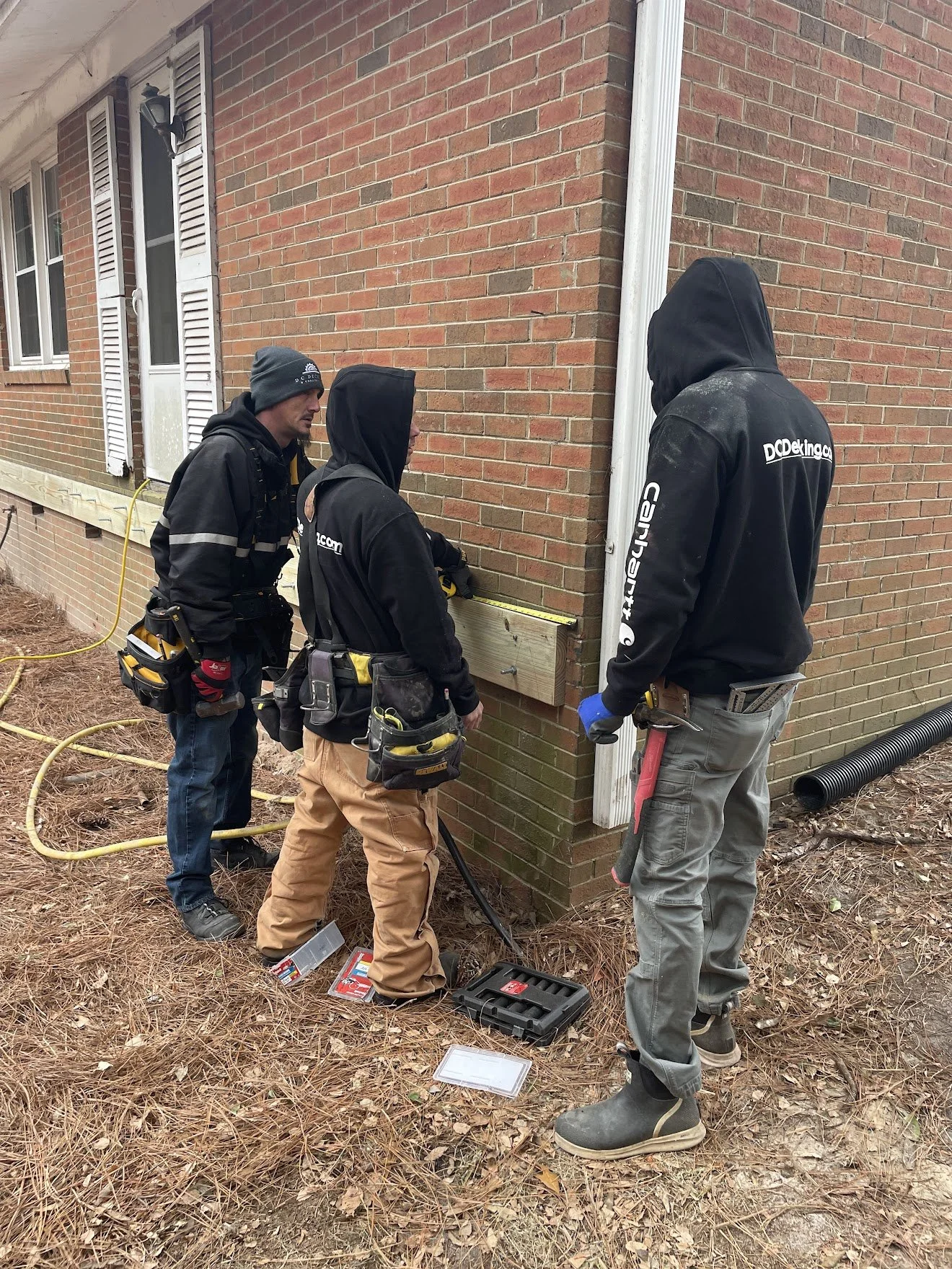 Three construction workers inspect the base of a brick house exterior, wearing safety gear and working with tools on the ground.