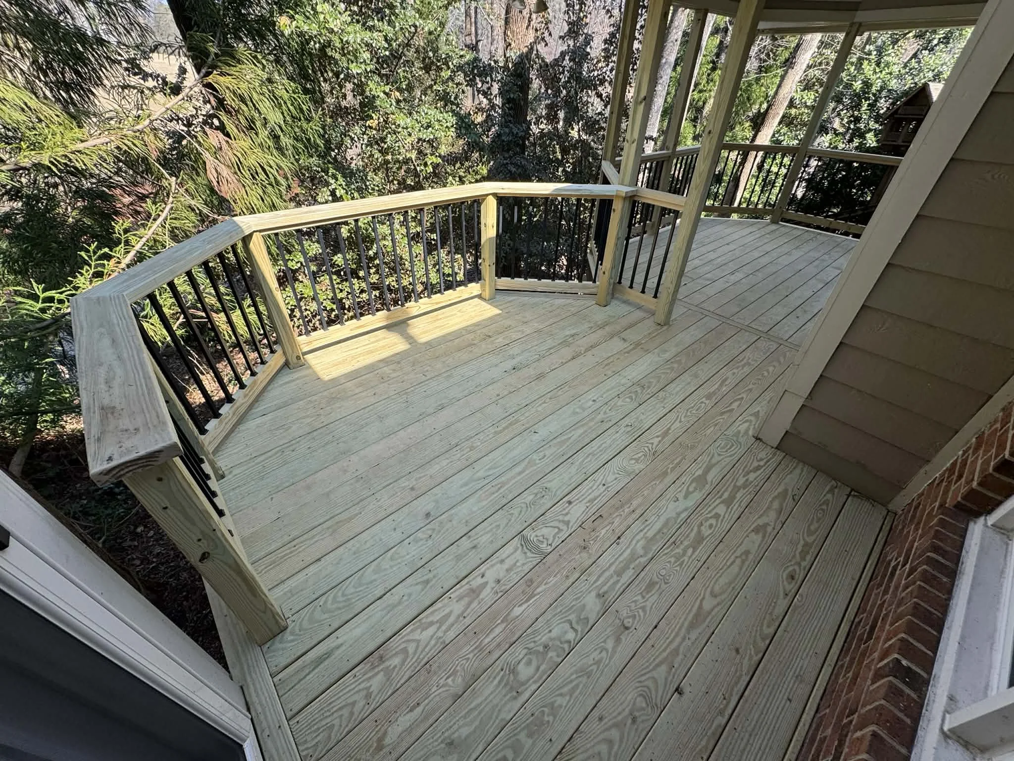Newly constructed wooden deck balcony with railing and stairs, attached to a house surrounded by trees and greenery.
