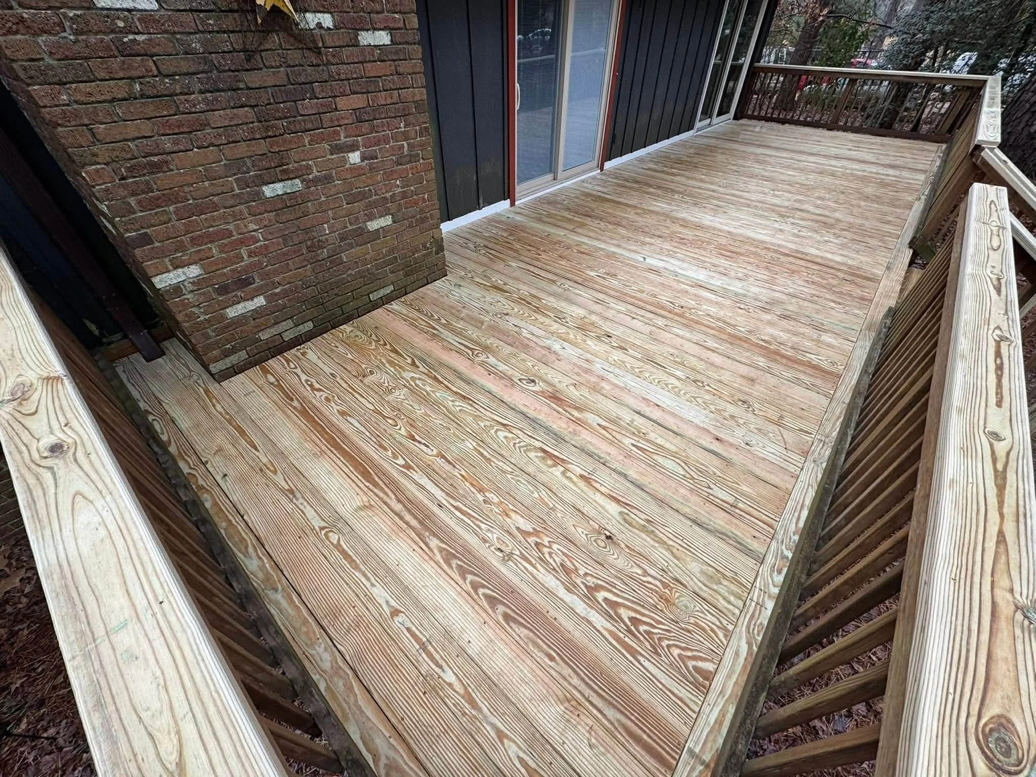A newly built wooden deck with light-colored wood planks and a railing, attached to a brick house with a sliding glass door and dark siding.