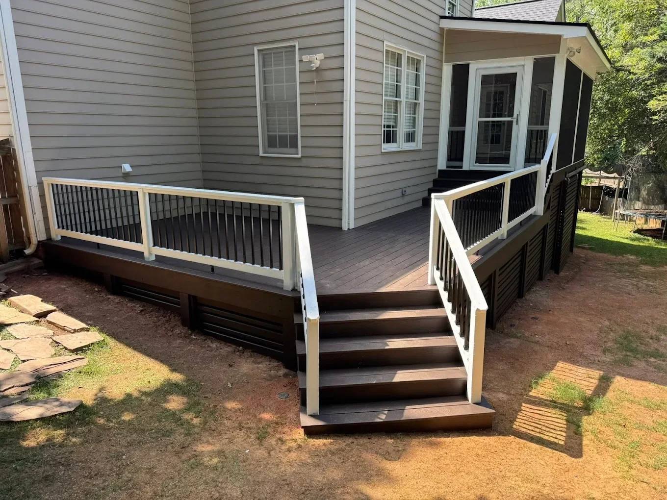 A raised wooden deck with stairs and white and black railings attached to the back of a house with beige siding and white-framed windows, in a backyard with grass and a trampoline.