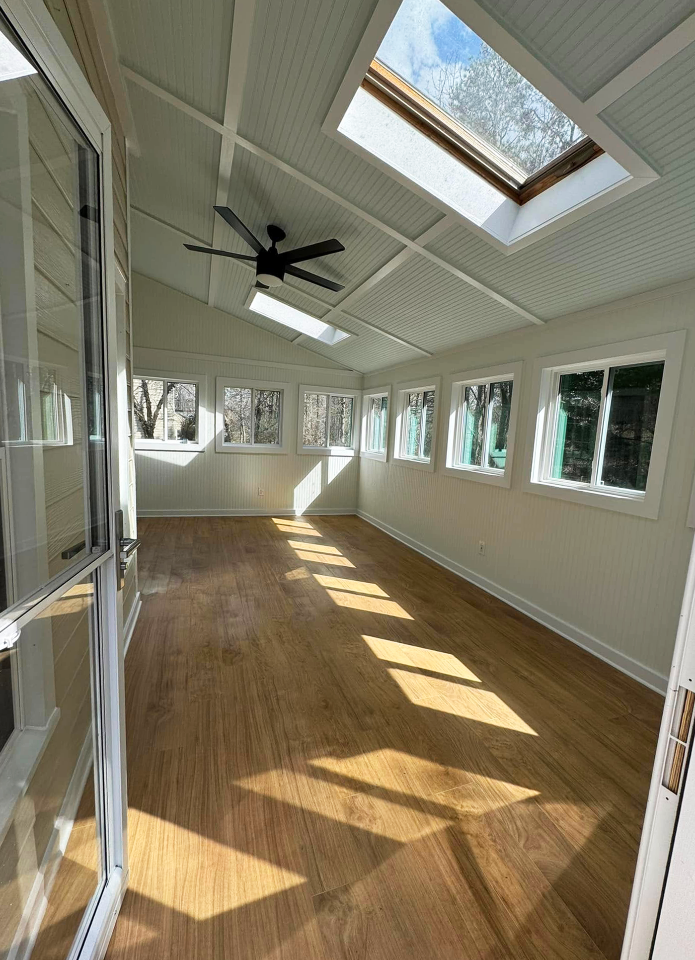 An enclosed porch or sunroom with multiple windows, a skylight, a ceiling fan, wood flooring, and sunlight casting shadows on the floor.