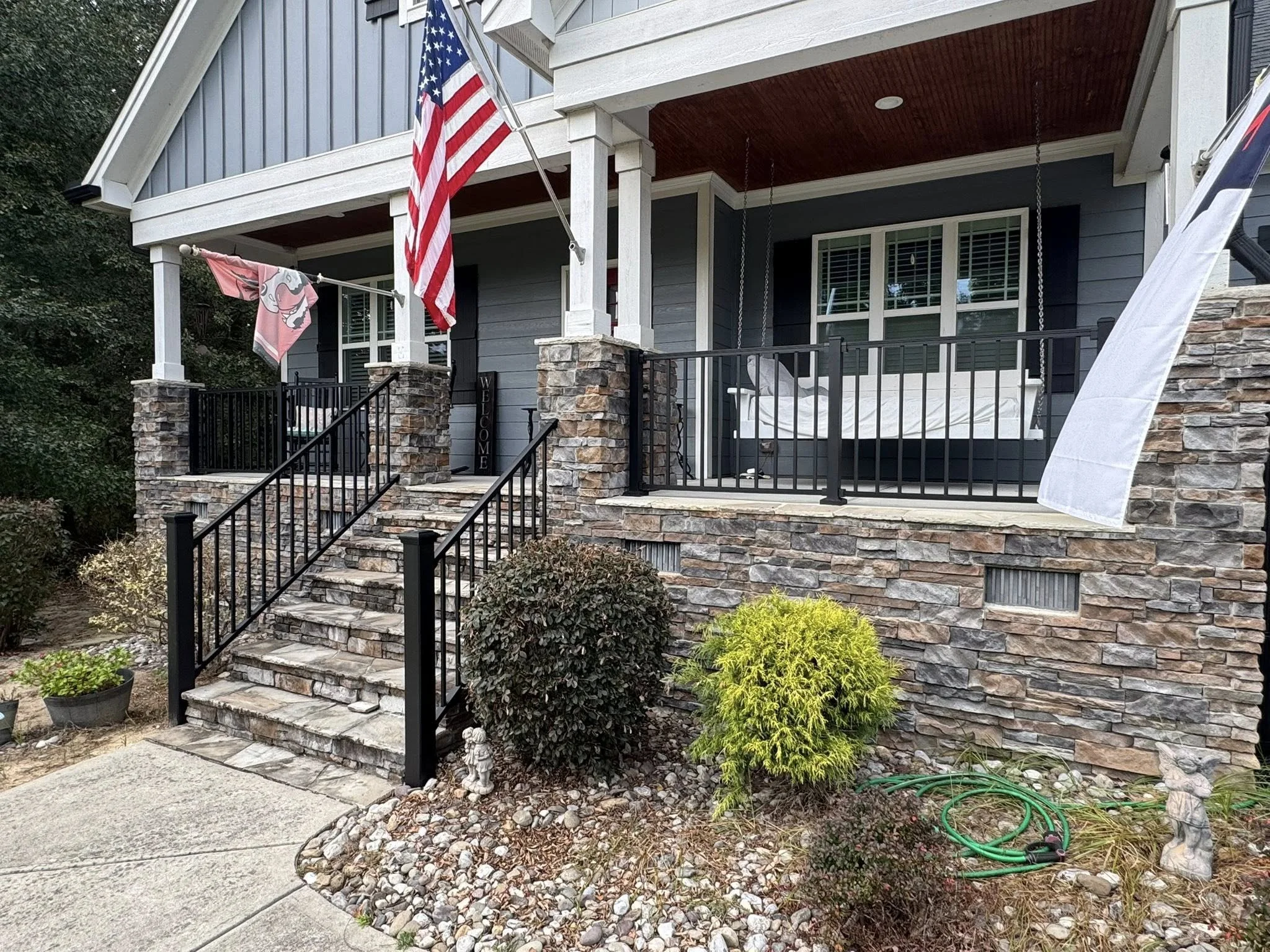 Front porch of a house with a stone staircase, black metal railing, and a swing. American flag and a pink towel hanging on the porch. Bushes and plants at the base of the stairs, garden hose on the ground, and decorative figurines.