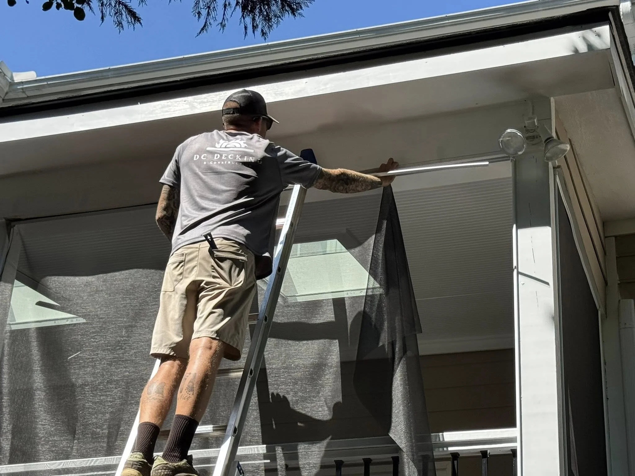 A man standing on a ladder installing or adjusting a screen or curtain on the exterior of a house or porch.
