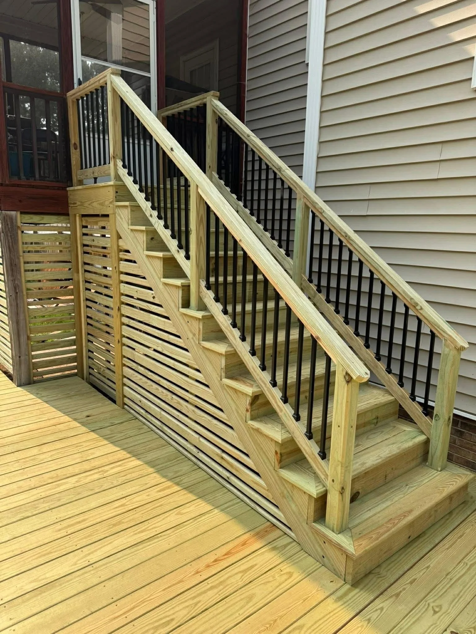 Newly built wooden outdoor staircase with black metal railing leading to a screened porch, attached to a beige siding house with a small set of steps at the bottom.